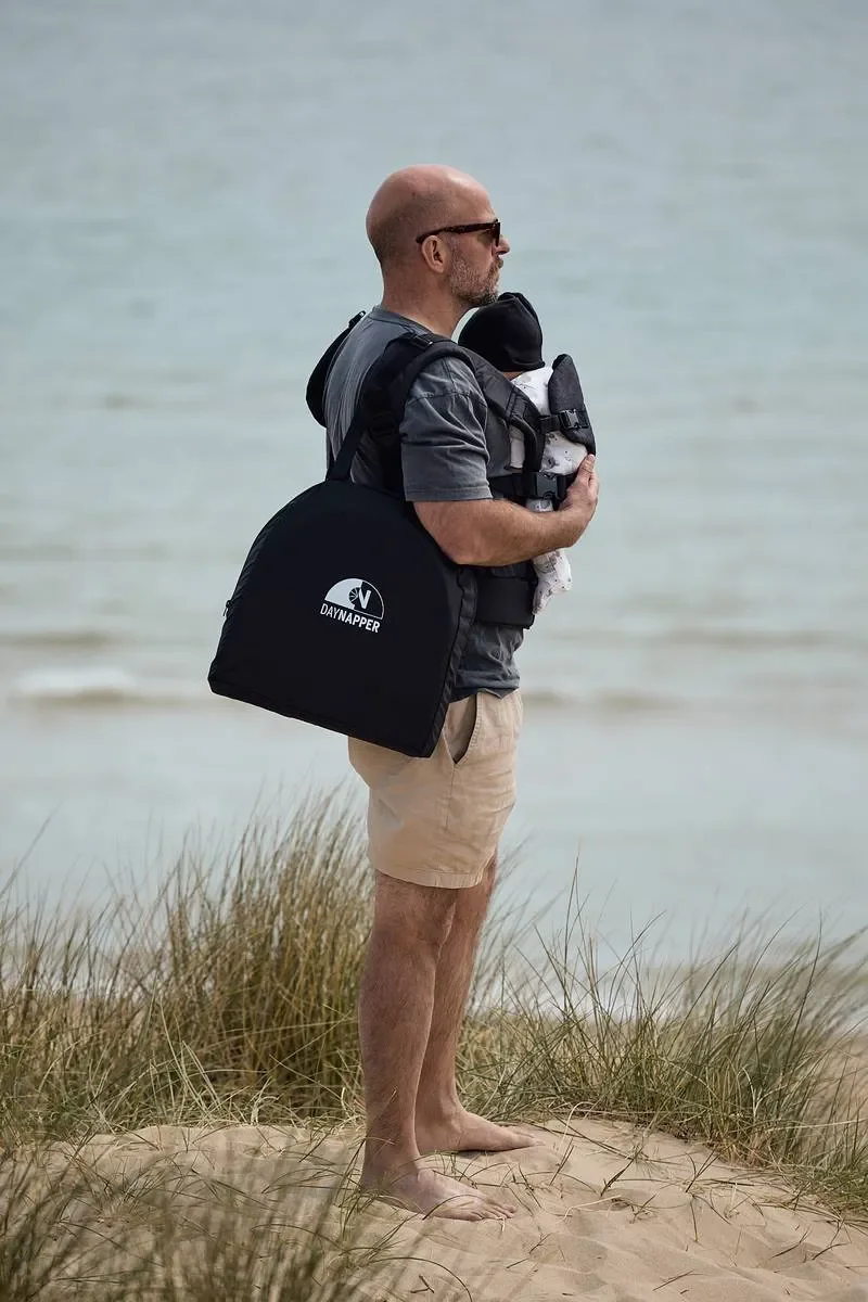 A father standing on a sandy beach with grass, holding a baby in a baby carrier, wearing sunglasses and shorts, with the ocean in the background and DayNapper over his shoulder.