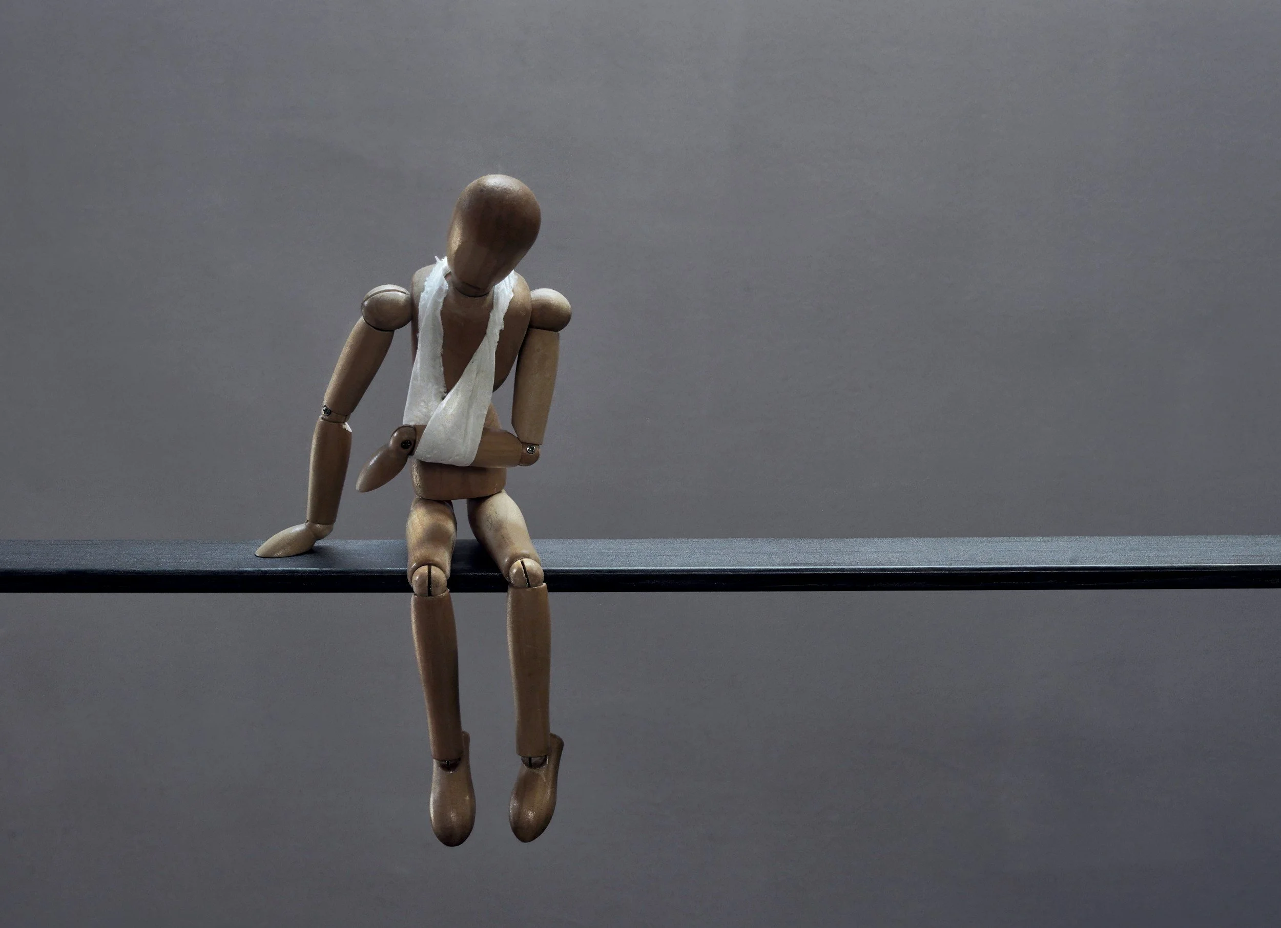 A wooden artist mannequin sitting on a dark surface with a gray background, wearing a white bandage around its neck and chest area.