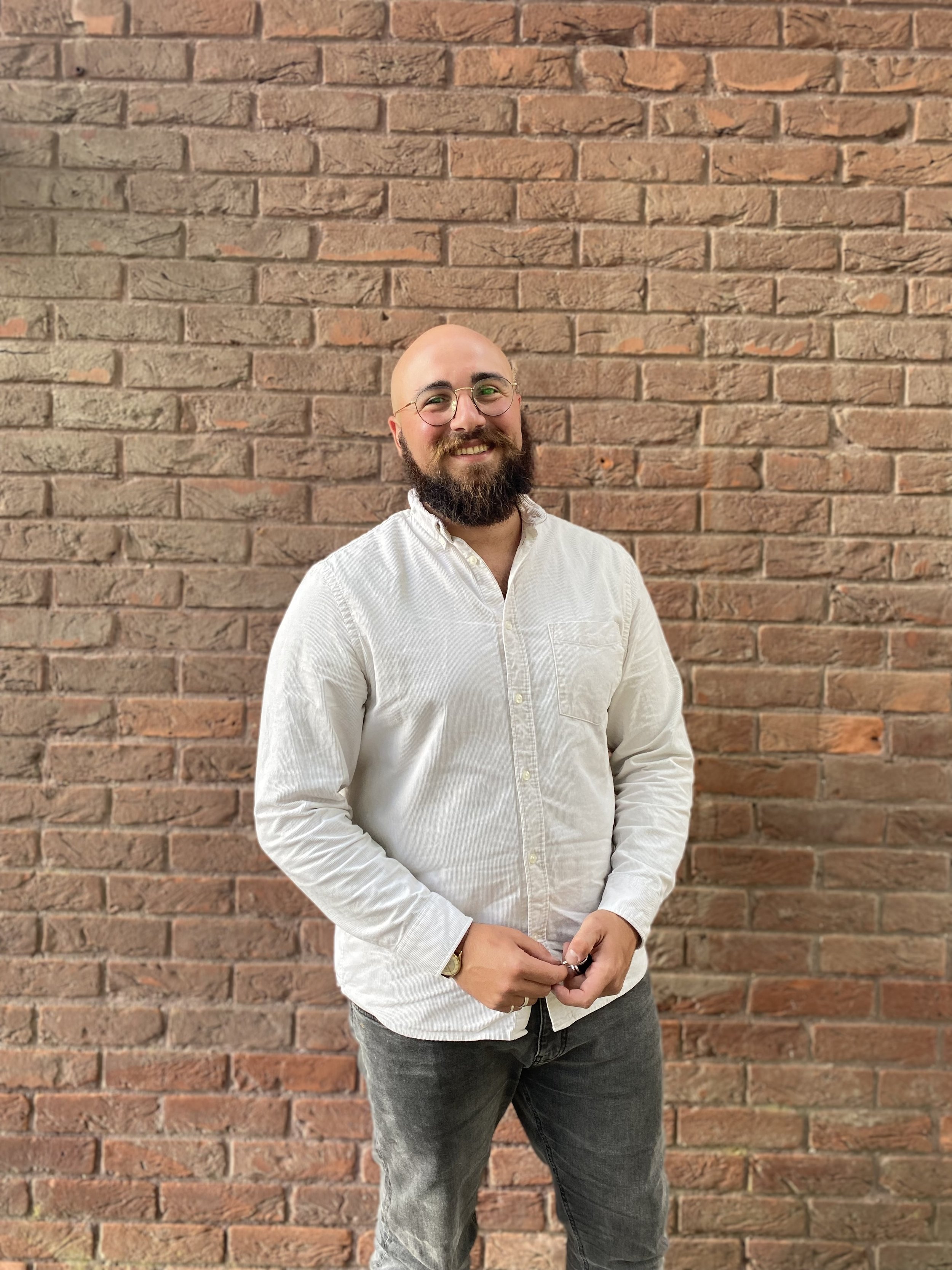 A smiling man with a beard, glasses, and a bald head standing in front of a brick wall. He is wearing a white button-up shirt and dark jeans, holding something in his hands.