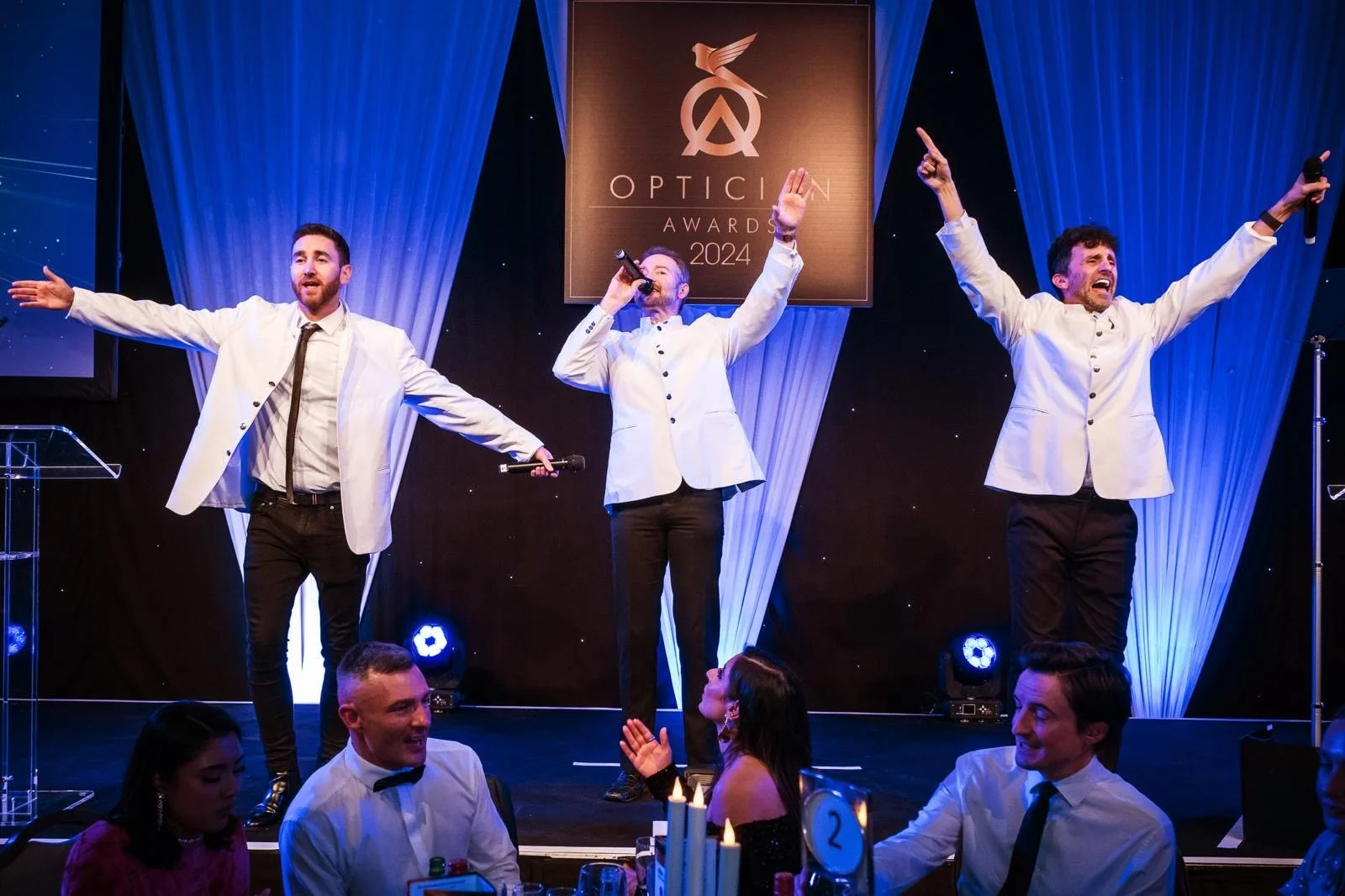 Three men in white jackets and black pants are on stage at the Opticin Awards 2024, singing or speaking into microphones with their arms raised, while an audience sits at tables below.