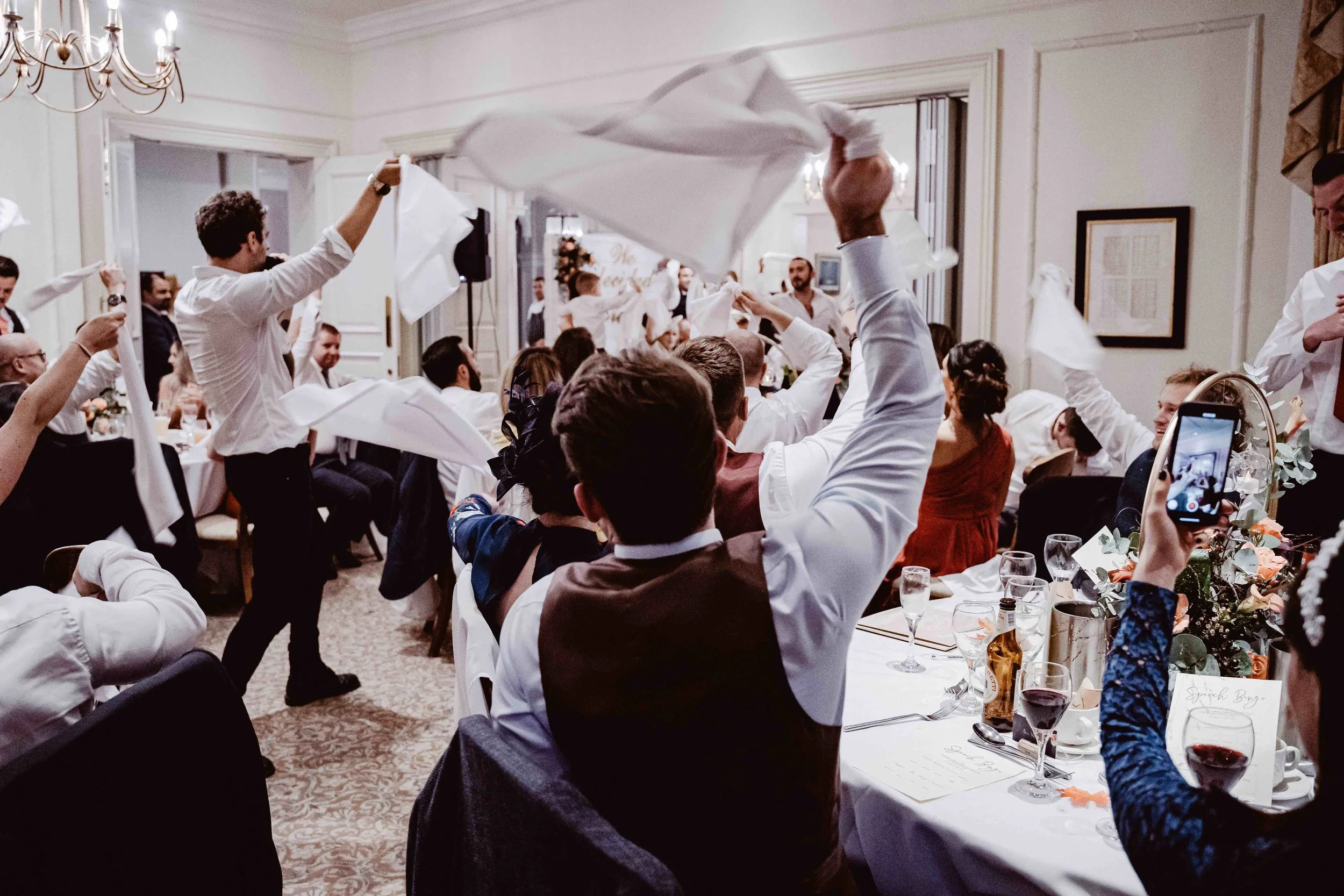 Guests celebrating at a wedding reception with people dancing and waving white napkins.