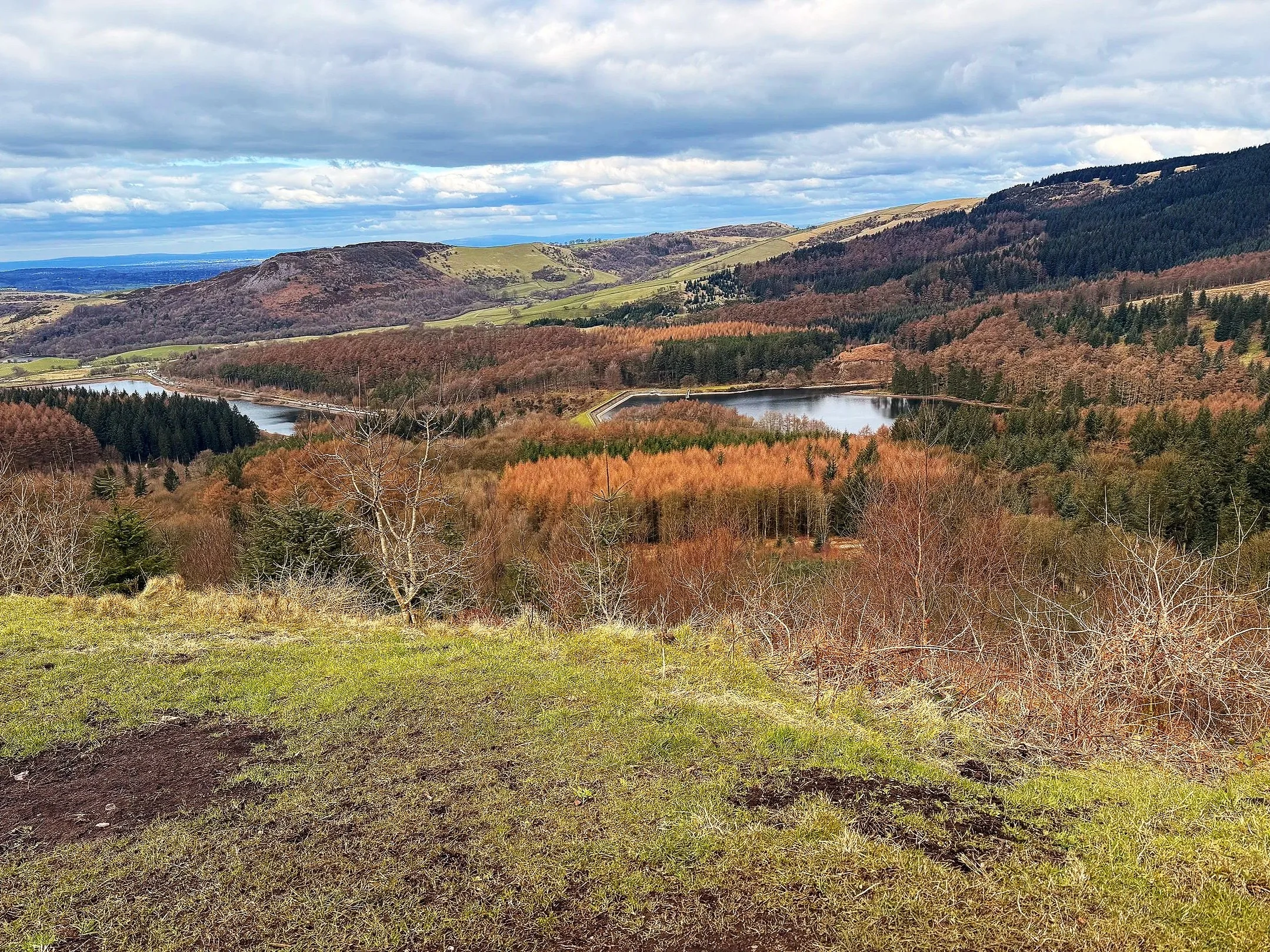 Scenic landscape with rolling hills, a lake, and autumn trees in a valley, under a partly cloudy sky.