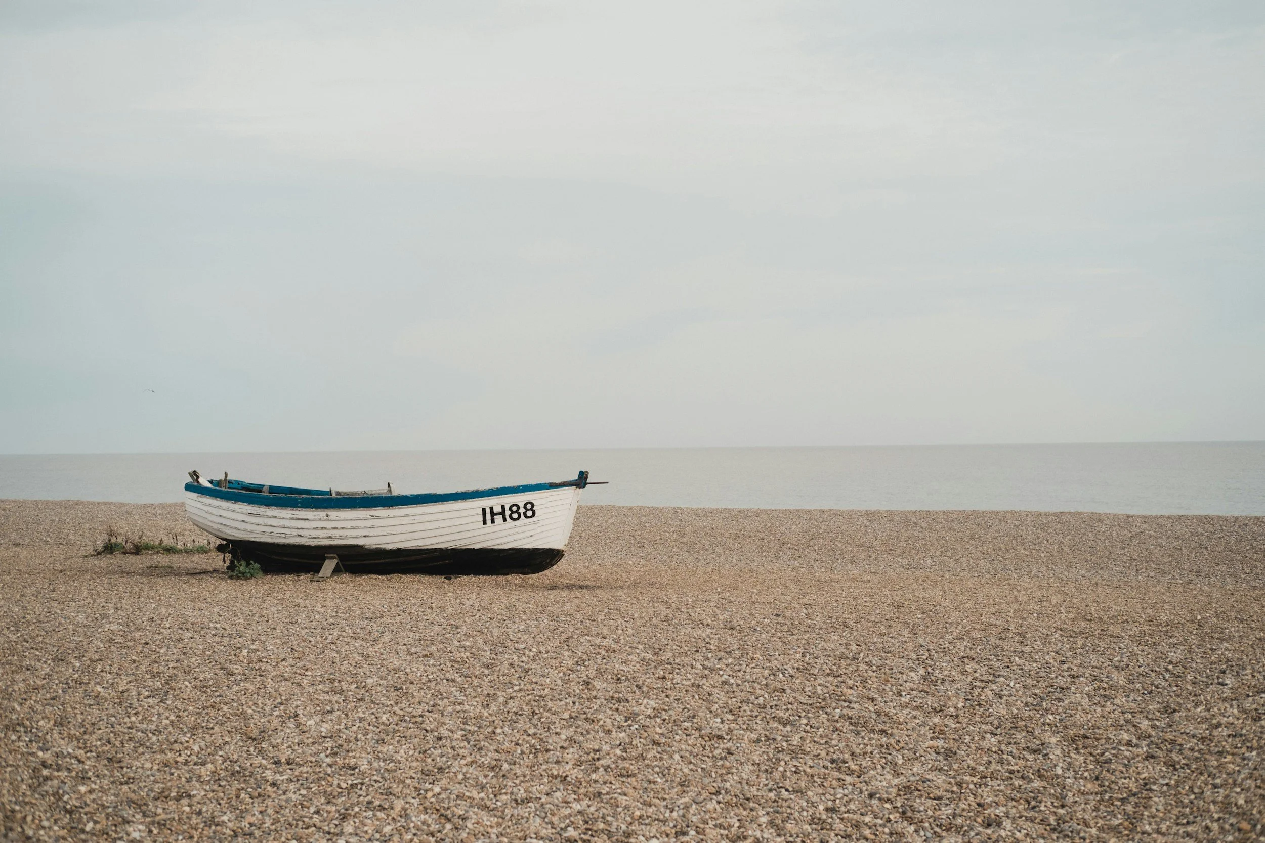 A white and black boat resting on a pebble beach near the water, with a calm sea and an overcast sky in the background.