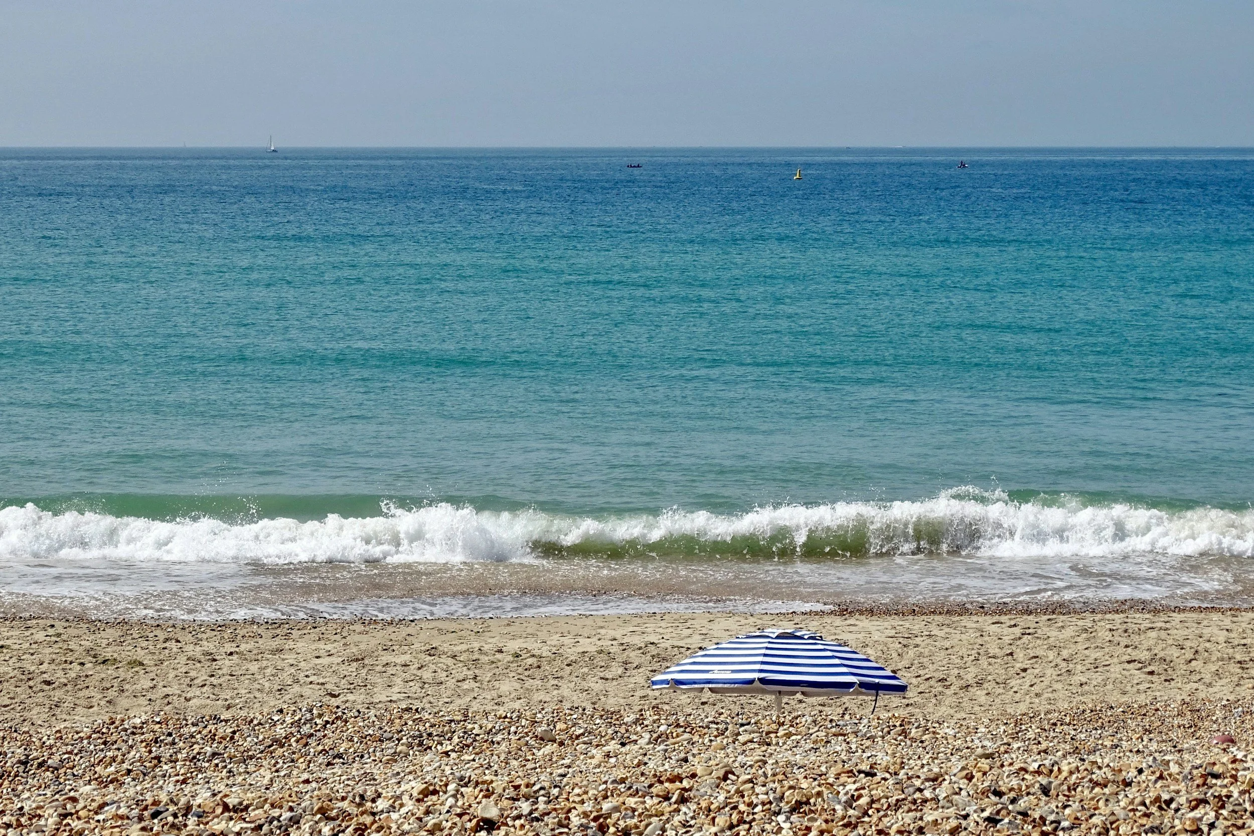Beach scene with a striped blue and white umbrella on rocky shore, gentle waves, and the ocean in the background with a few small boats.
