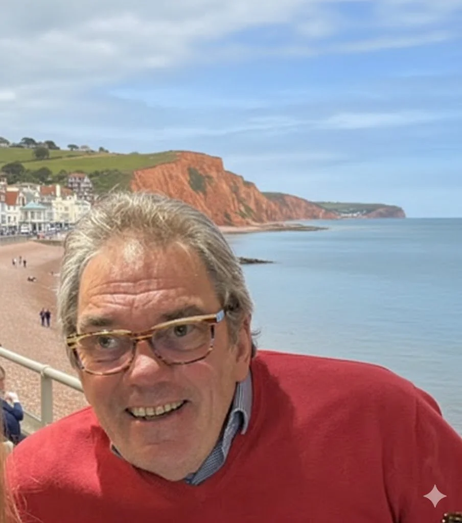 A smiling man with gray hair and glasses, wearing a red sweater, standing near a seaside with cliffs and houses in the background.
