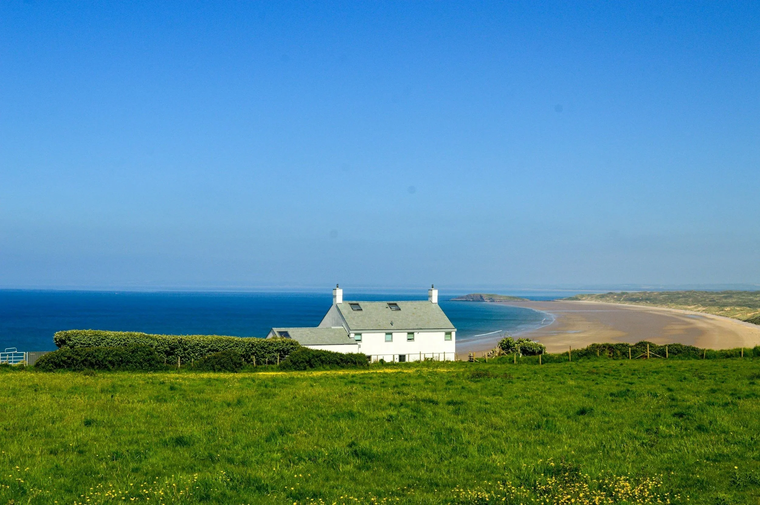 A white house with a gray roof near a green field, overlooking a sandy beach and the ocean under a blue sky.