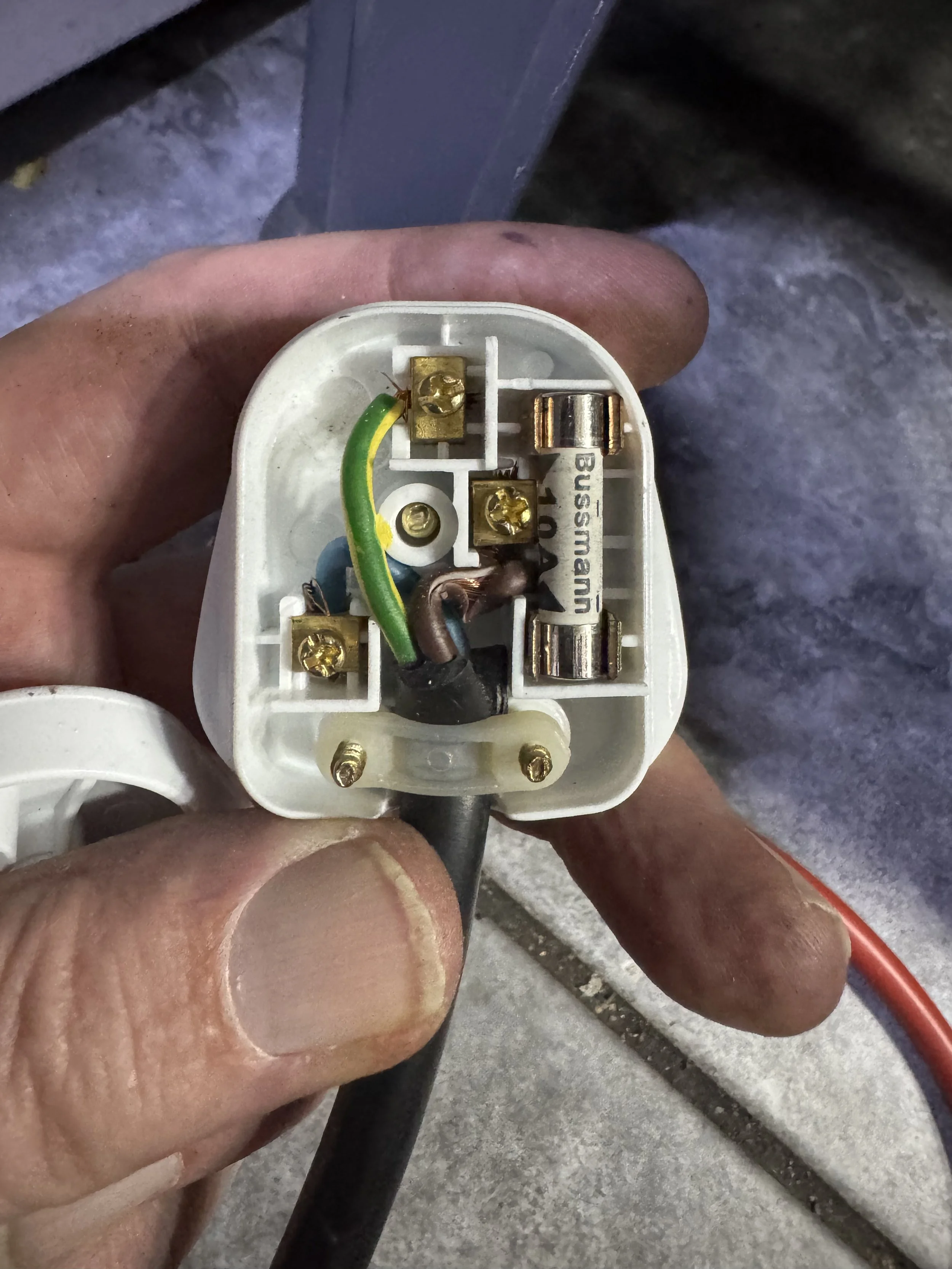 Close-up of an electrical plug with exposed wiring, showing gold screws, green/yellow grounding wire, brown wire, and a white terminal block, held by a person's hand.