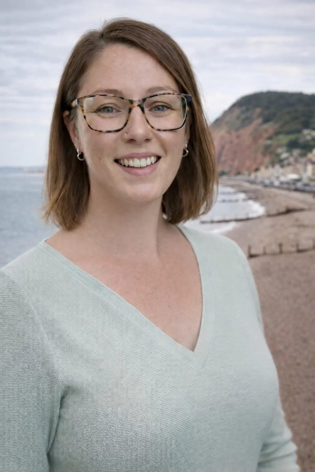 A smiling woman with glasses and earrings standing outdoors near the beach with cliffs and ocean in the background.