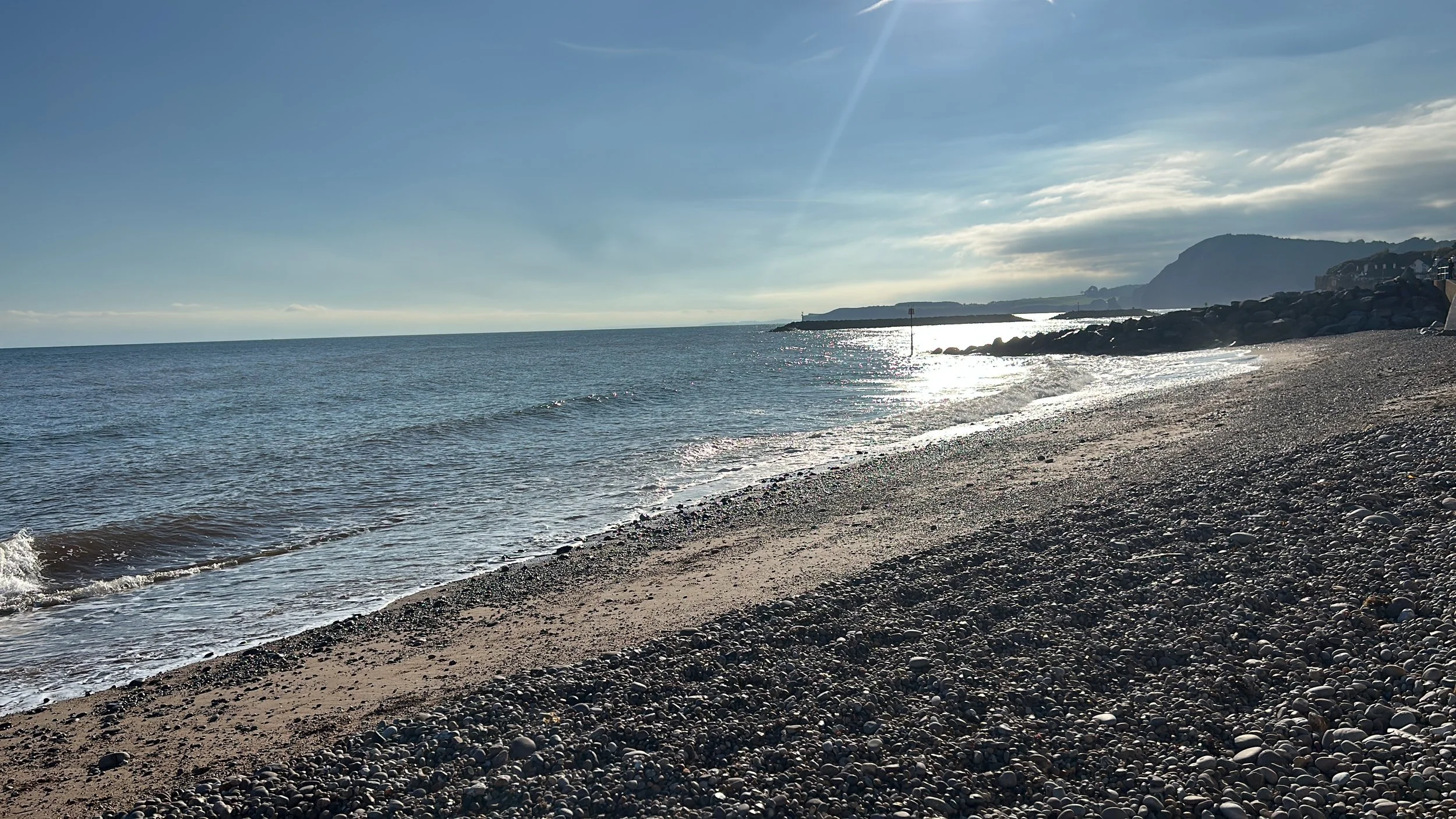 Sunlit beach with pebble shoreline, calm sea, and distant cliffs under a partly cloudy sky.