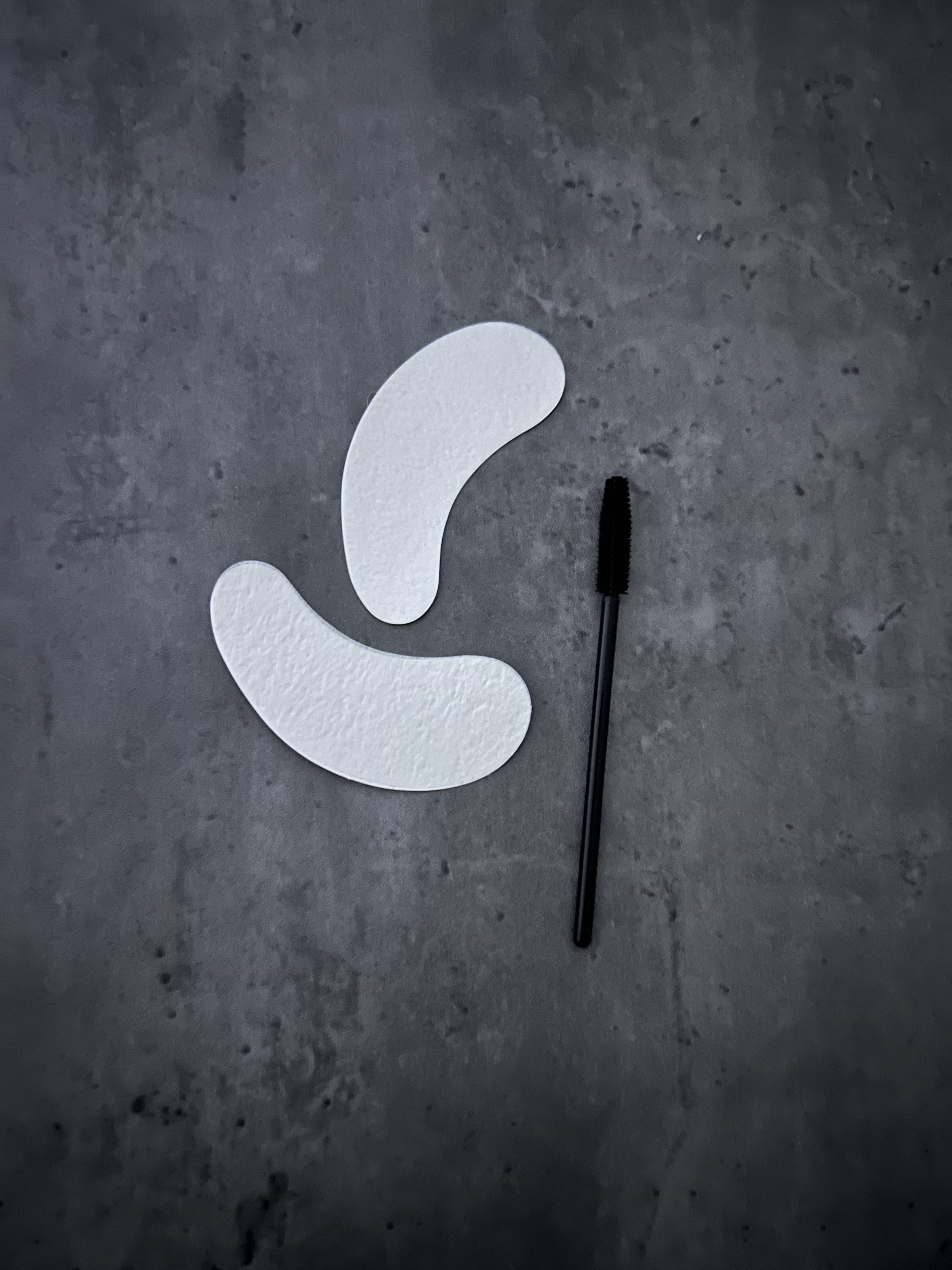 Two white paper patches, a mascara spoolie brush, all placed on a gray textured surface.