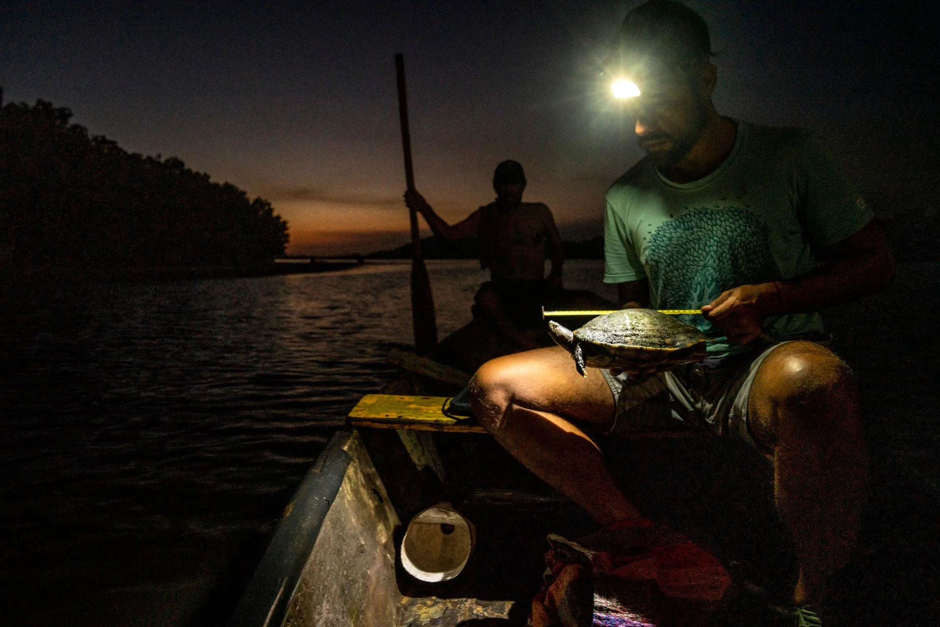 A man sitting on a small boat at dusk, holding a fish, with a paddle in the background, and another person in the distance. The sky is darkening with the last light of sunset, and a bright headlamp is on the man's forehead.