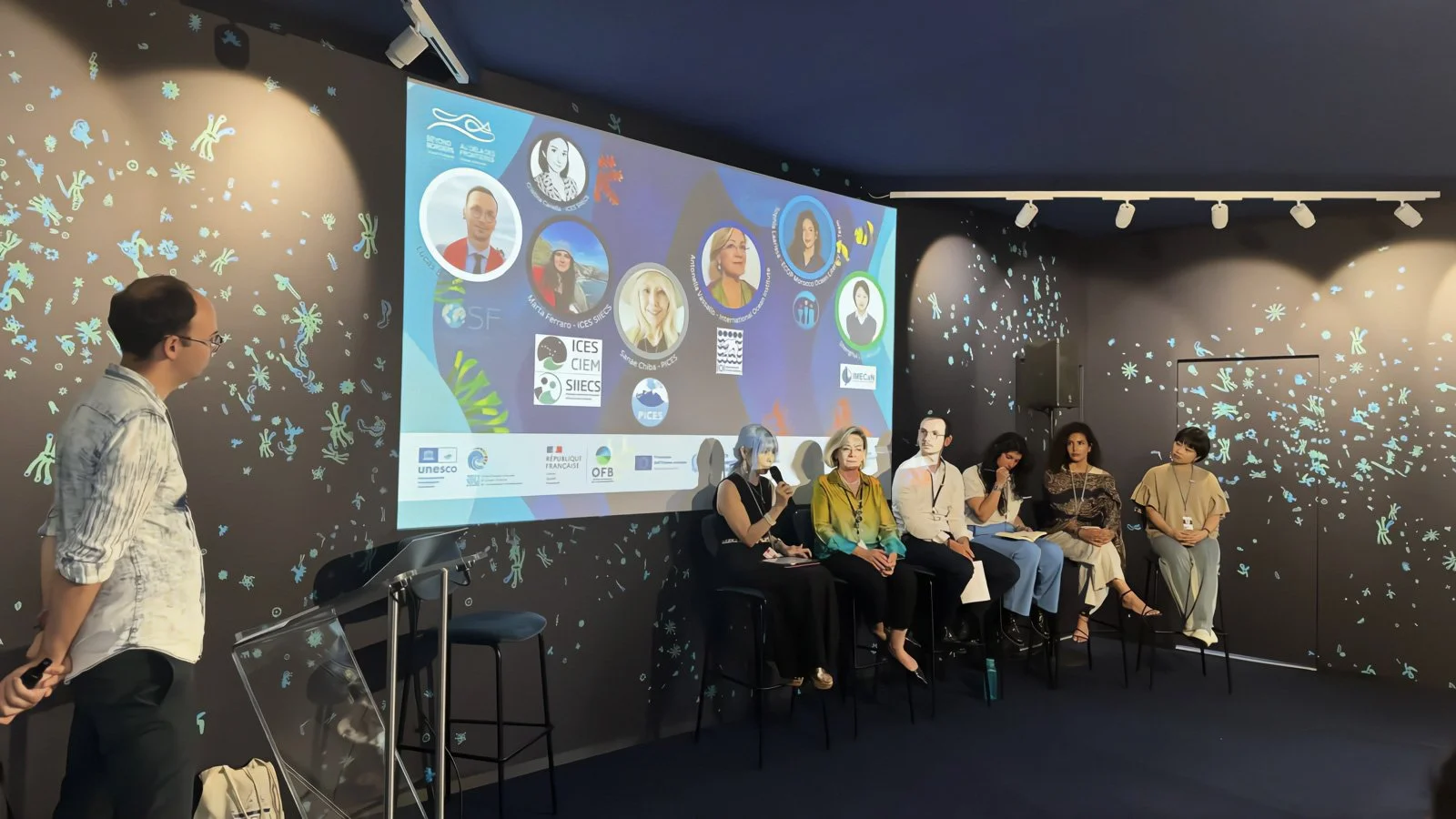 Panel discussion at a conference with six seated speakers on a stage, and a man standing to the left, looking at the screen. The backdrop features scientific illustrations of microbes and logos of organizations including UNESCO and European Union.