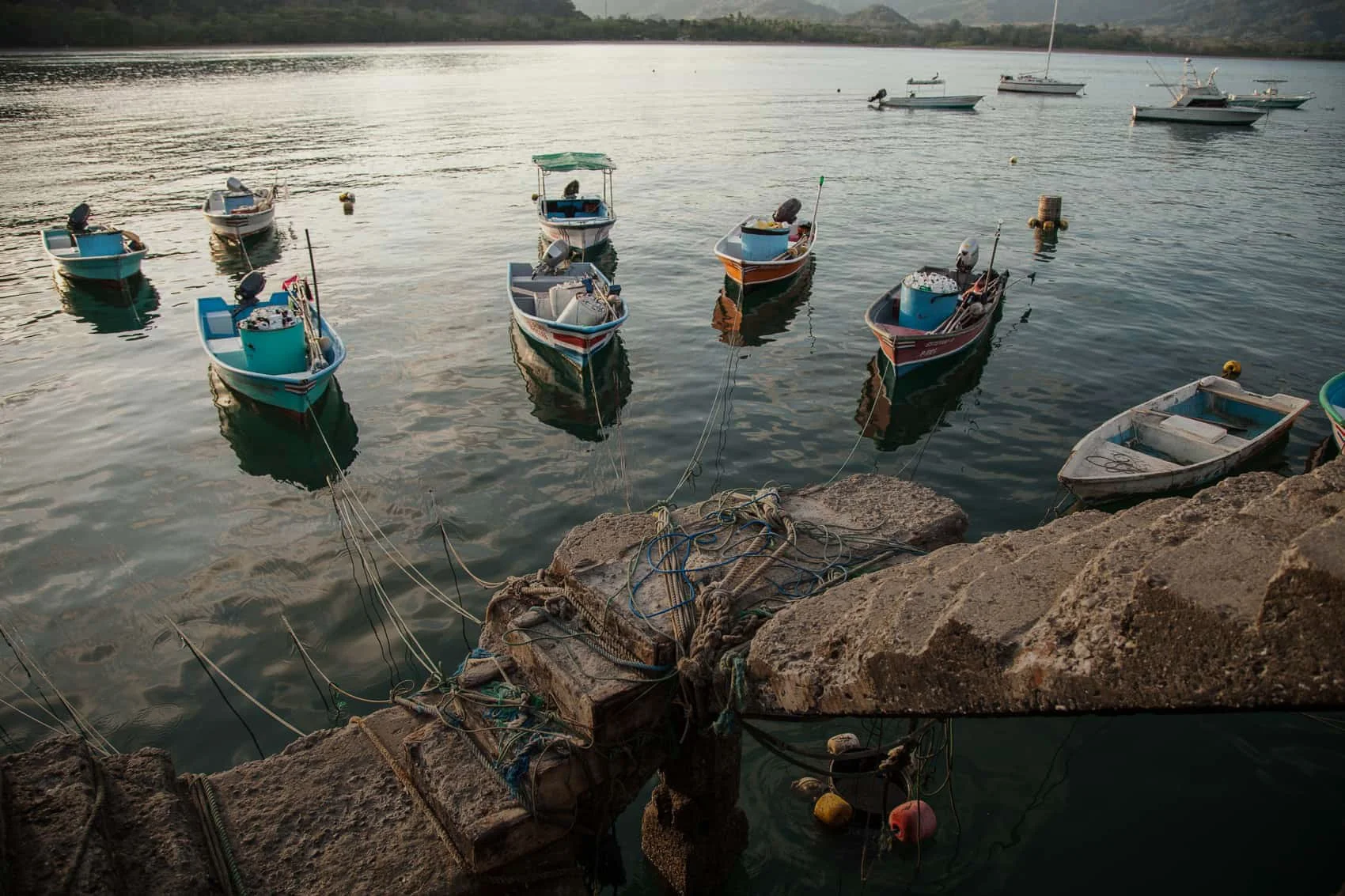 Small boats tied to a stone dock on a calm body of water with distant hills in the background.