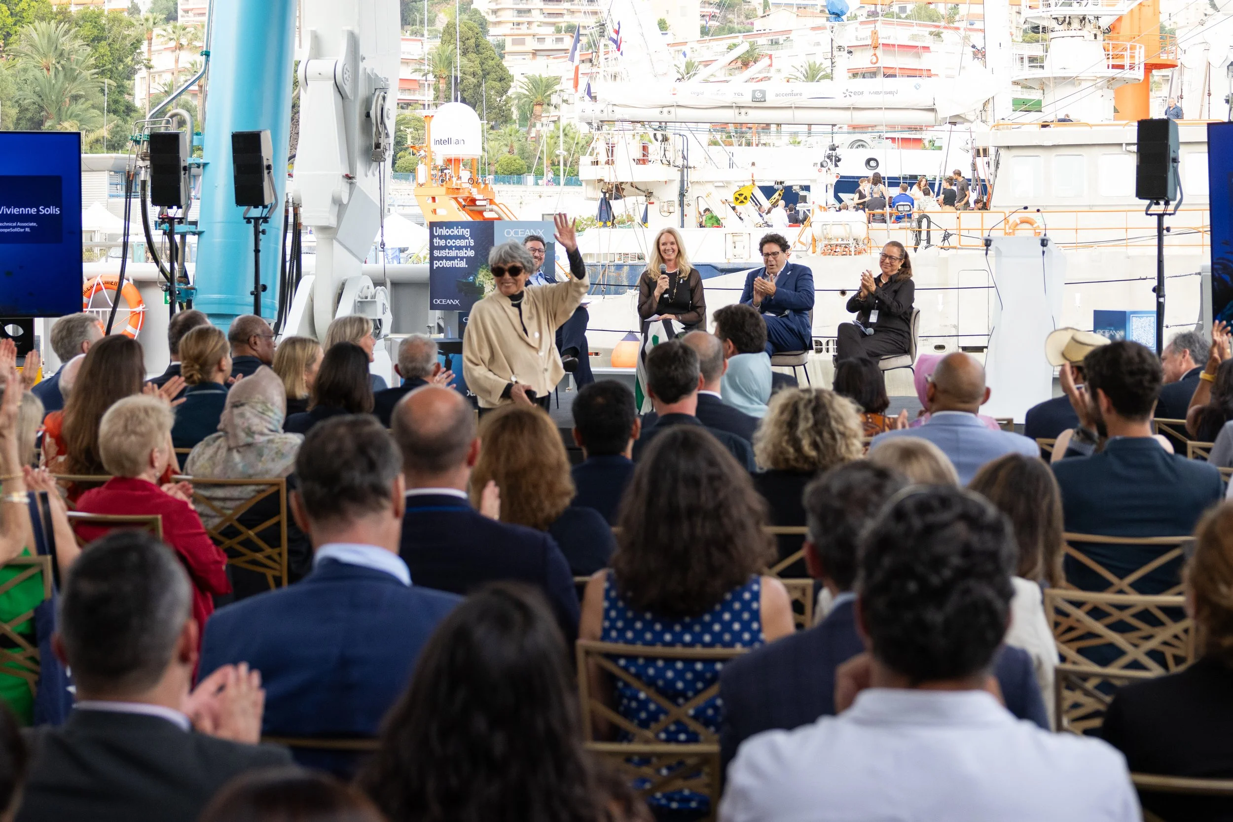 People attending a conference outdoors near boats, with a woman standing and speaking into a microphone, and several people seated and clapping, in front of large ships at a marina.