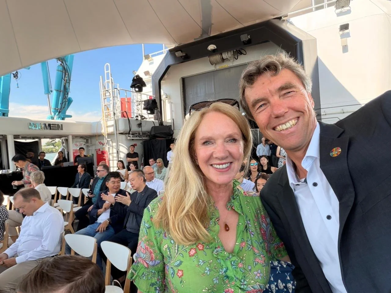 Two smiling people taking a selfie at an outdoor event with many seated attendees and a bright sky in the background.