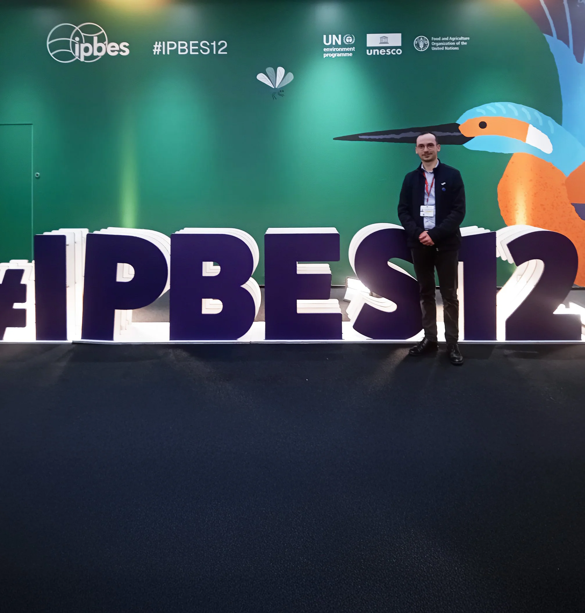 A man stands in front of a green wall with logos of IPBES, UN environment programme, UNESCO, and FAO. There is a colorful illustration of a bird on the wall. The man is wearing glasses, a black jacket, and a lanyard.