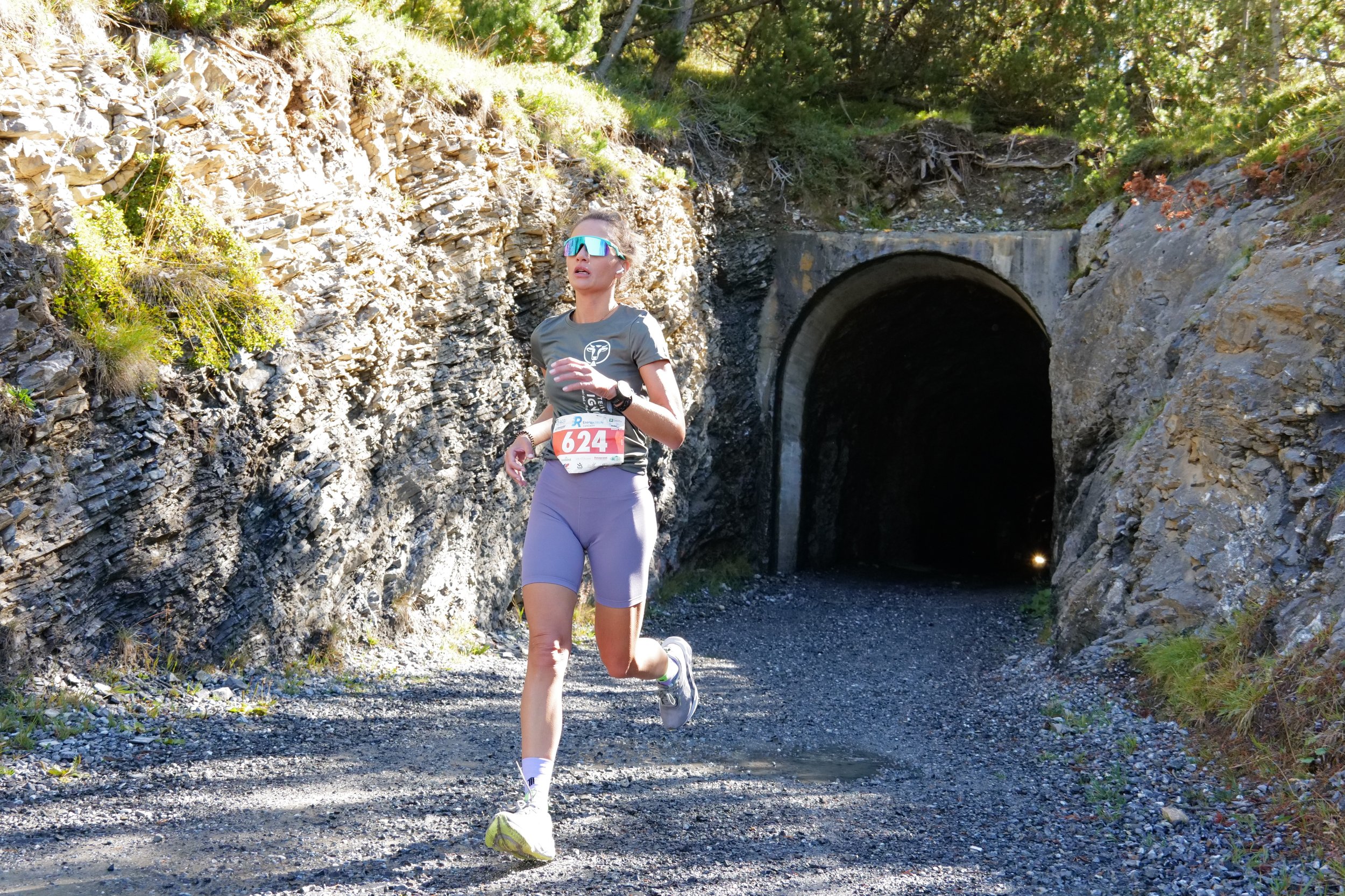 Una donna corre lungo un sentiero in un'area montana, con un tunnel in pietra sullo sfondo, indossando abbigliamento da corsa e occhiali da sole