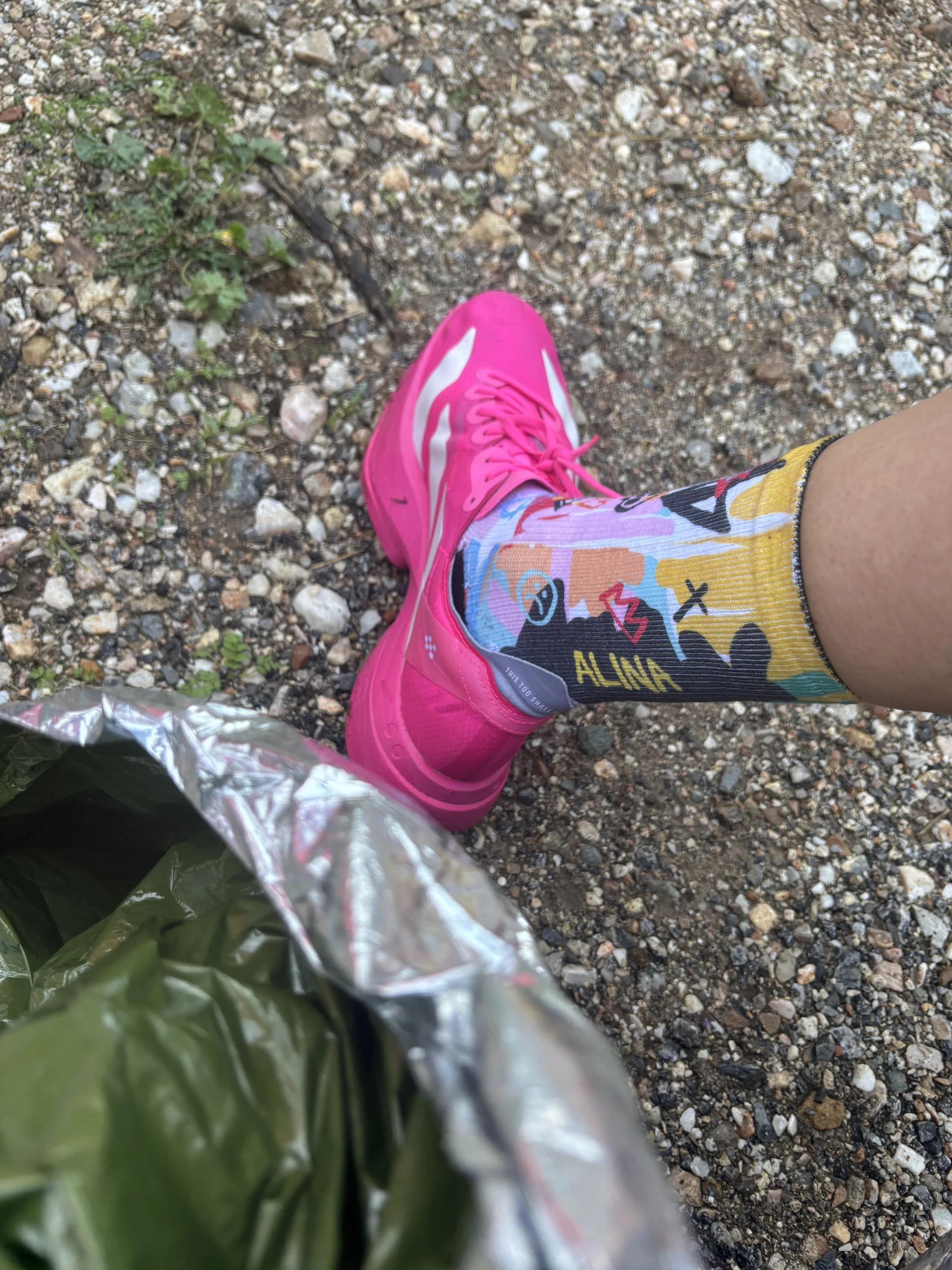 Scarpe rosa con calzini colorati, sulla ghiaia di un sentiero, con in mano un sacchetto di plastica verde.