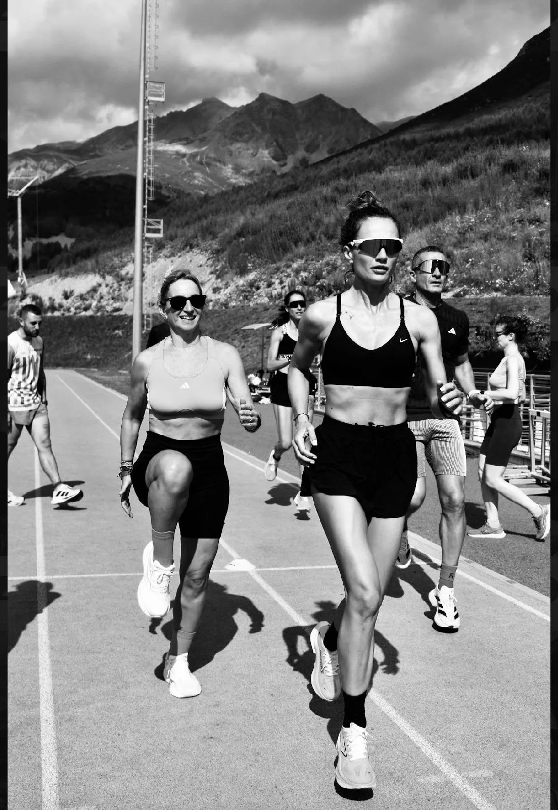 Gruppo di persone che corre all'aperto su una pista, con montagne sullo sfondo, in una giornata nuvolosa, vista in bianco e nero.