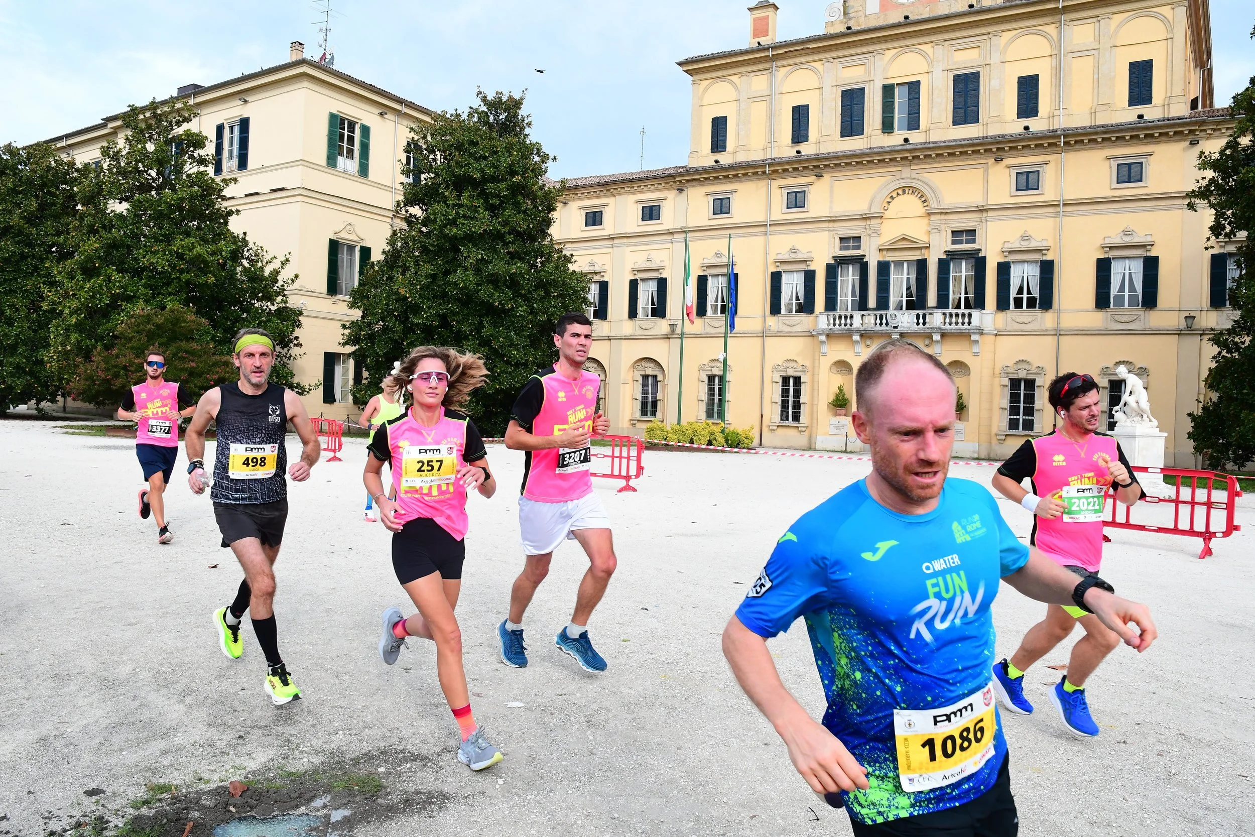 Gruppo di persone che corre durante una maratona davanti a un edificio storico con bandiere italiane ed europee alcune piante e statue.