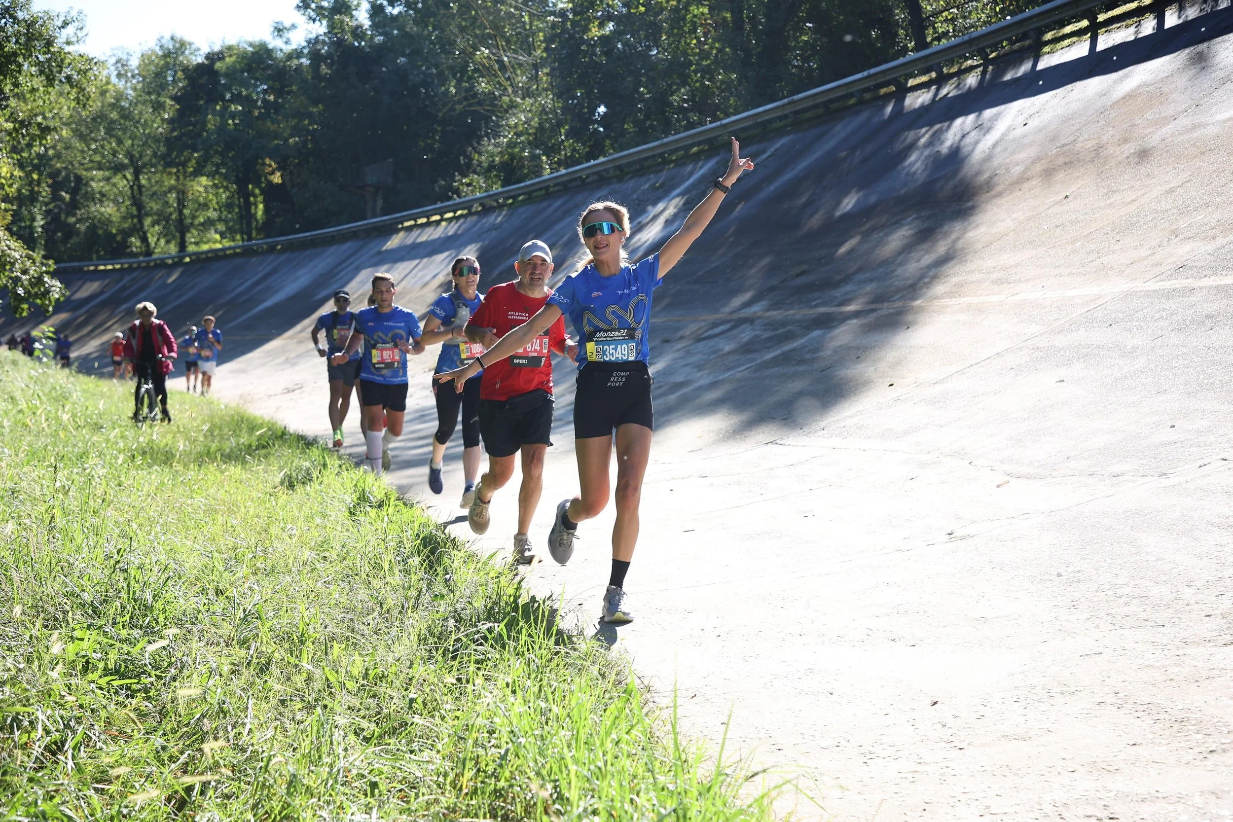 Gruppo di persone che corre su un sentiero naturale accanto a un grande muro inclinato, con alberi e vegetazione sullo sfondo, durante una competizione o maratona.