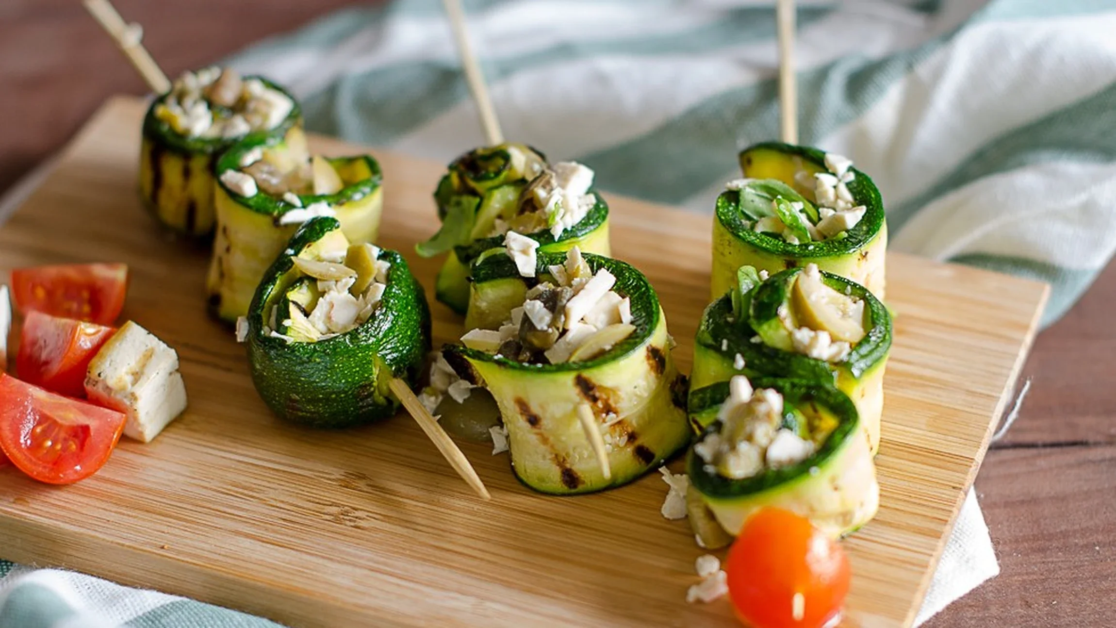 Involtini di zucchine ripieni di formaggio e noci, serviti su un tagliere di legno con pomodorini e formaggio accanto.