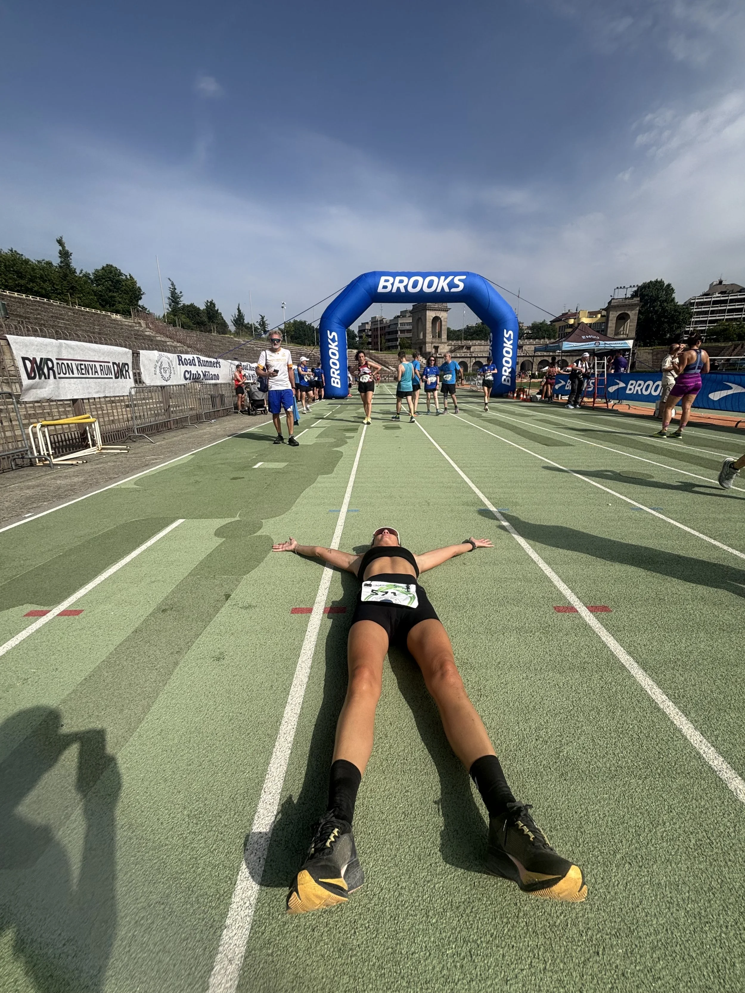 Atleta femminile sdraiata sul terreno di una pista di corsa dopo una gara, con arco pubblicitario blu sullo sfondo e altri partecipanti intorno, in una giornata di sole.