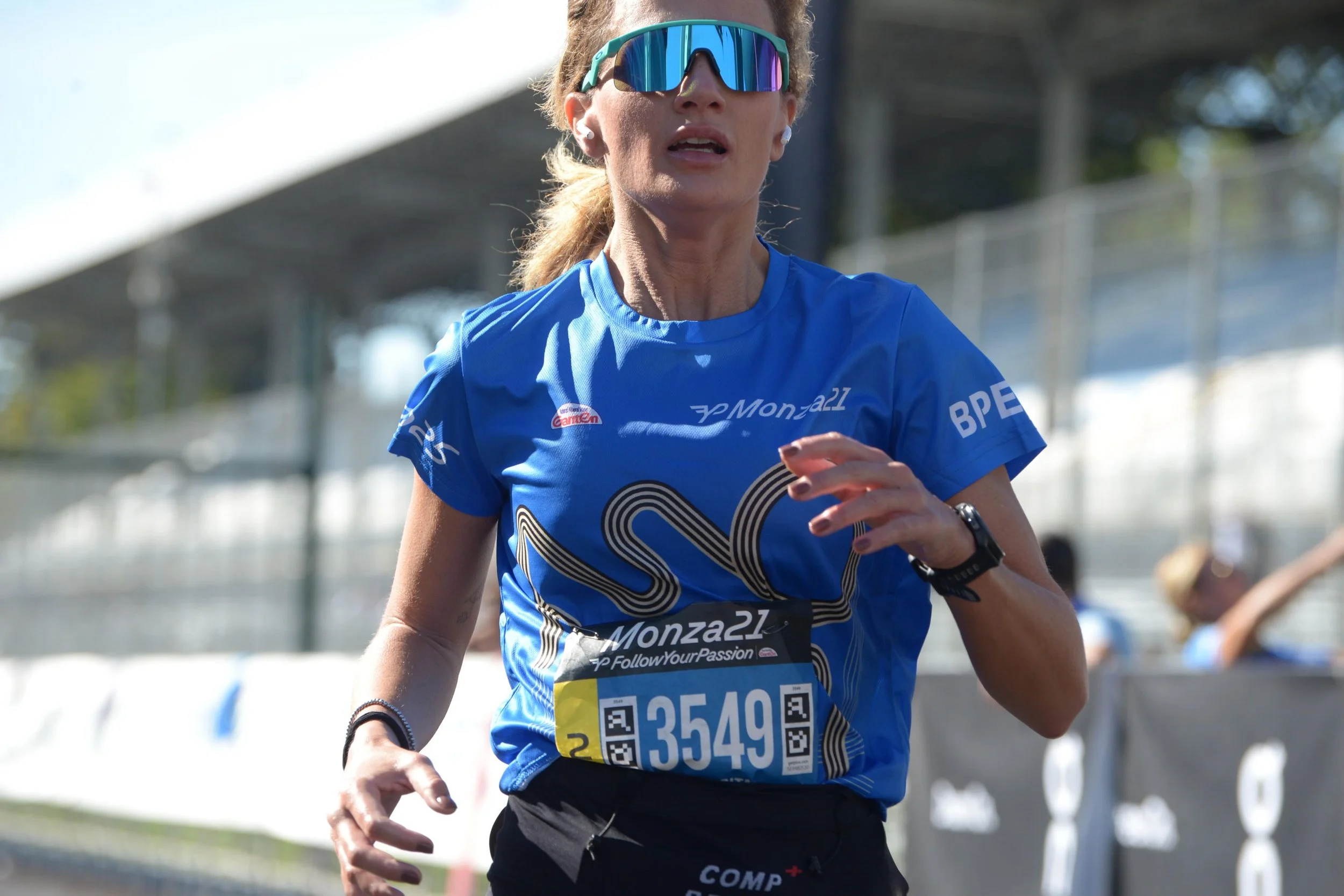 Una donna corre durante una maratona indossando un'abbigliamento da corsa blu e occhiali sportivi riflettenti, con un numero di gara 3549, in un ambiente all'aperto con strutture di gara sullo sfondo.