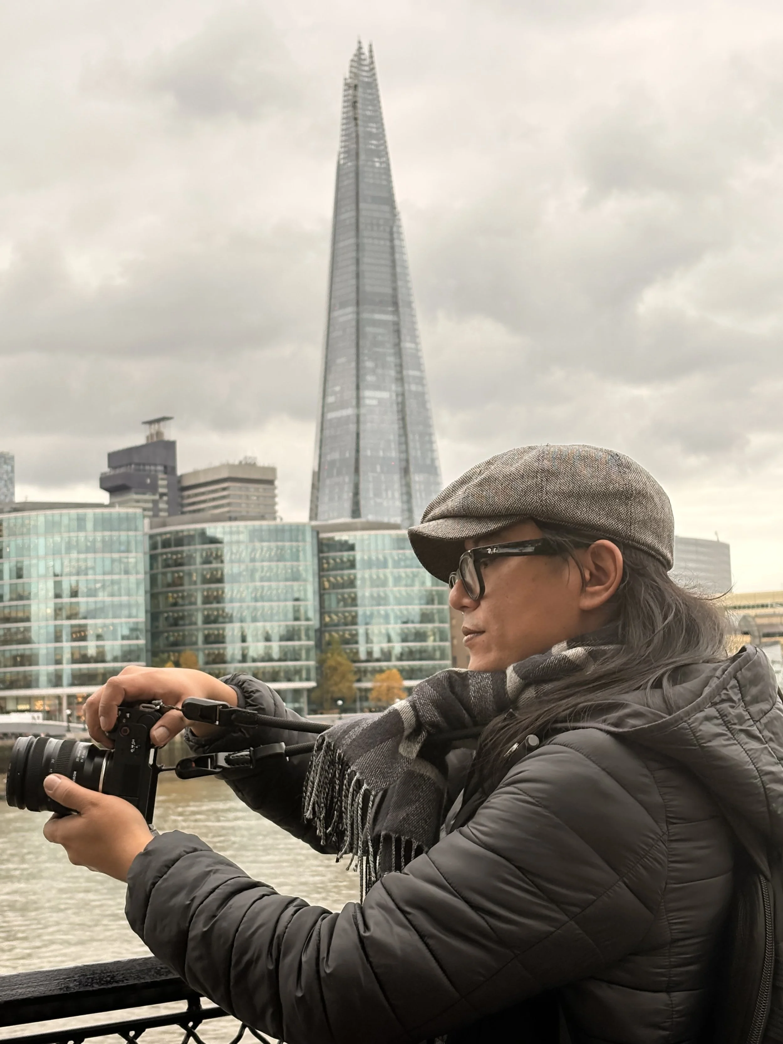 Pessoa com chapéu, óculos e casaco escuro segurando uma câmera de vídeo na beira do rio em Londres, com o edifício The Shard ao fundo.