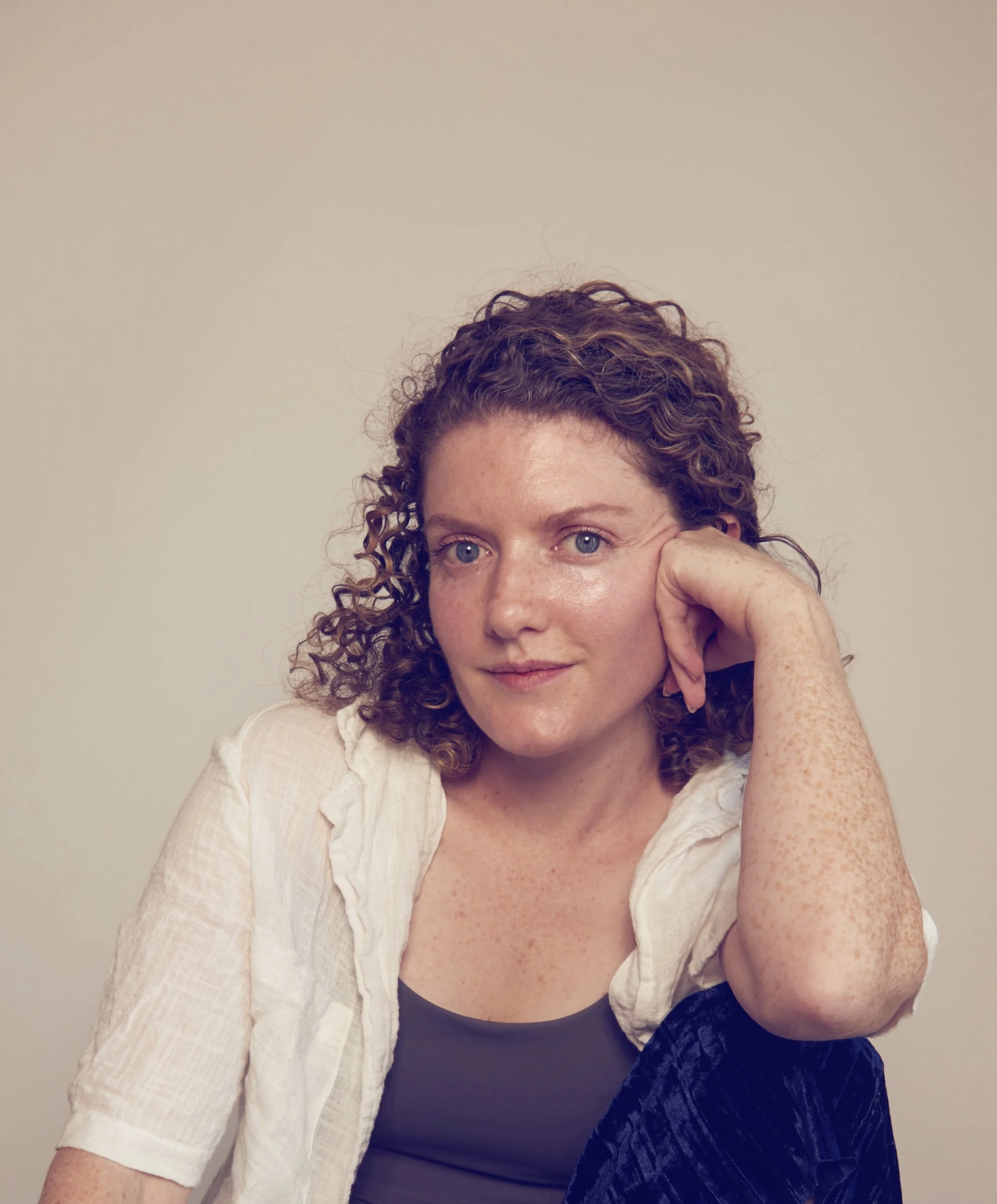 A woman with curly hair and blue eyes resting her chin on her hand, looking at the camera with a gentle smile, wearing a white textured jacket and a black top.