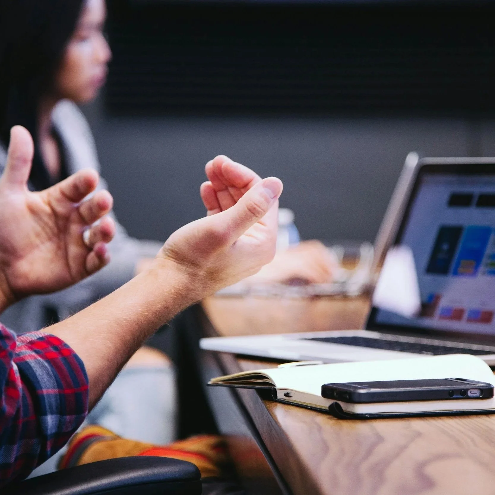 People in a meeting or discussion, with one person gesturing with their hand, a laptop, phone, notebook, and other office items on the table.