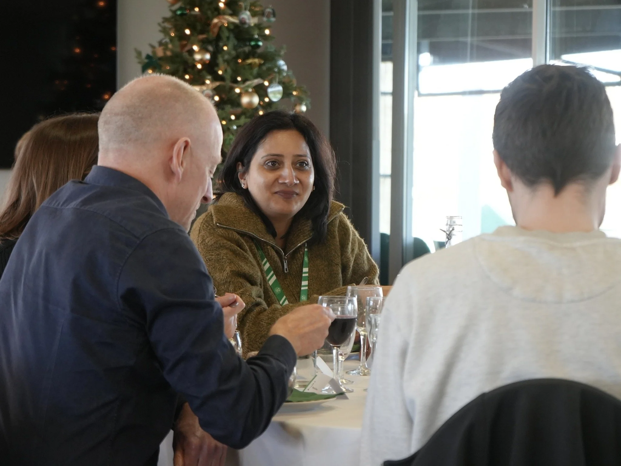 Four people sitting at a dining table decorated for Christmas, with a Christmas tree in the background, engaging in conversation and enjoying drinks.