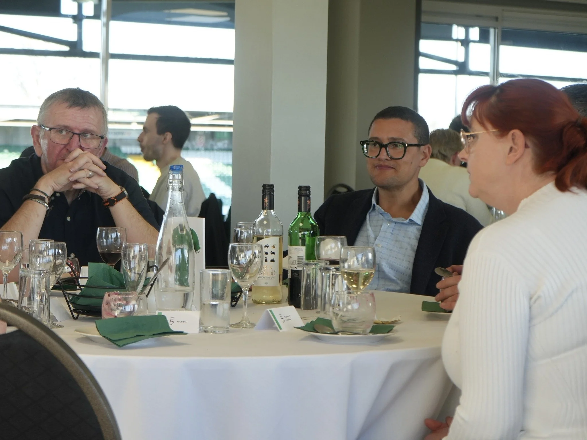 People seated at a dining table with wine bottles, glasses, and green napkins, engaged in conversation in a well-lit restaurant or event space.