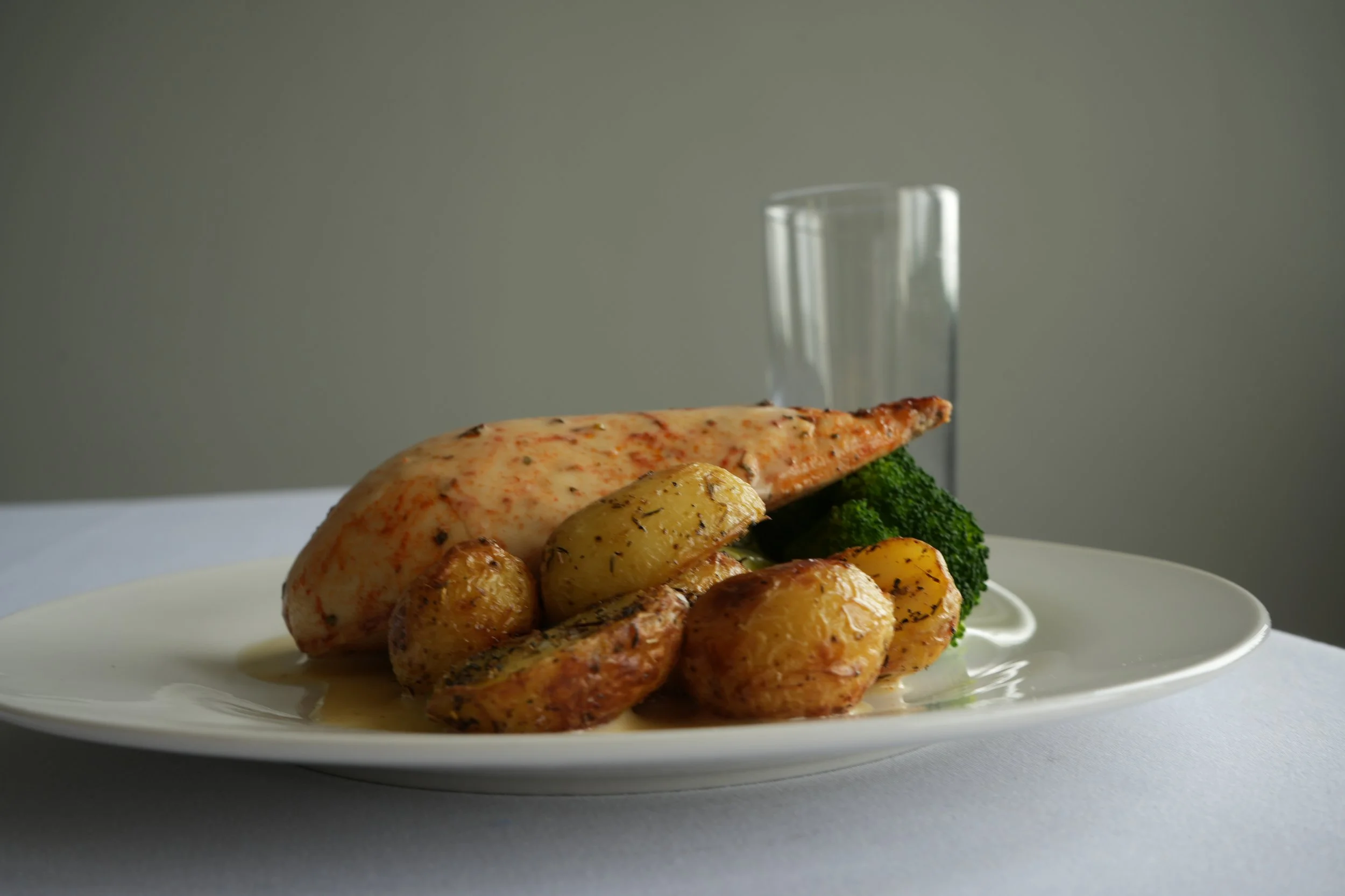 A plated meal featuring a piece of cooked fish fillet, roasted potatoes, broccoli, and a glass of water in the background.