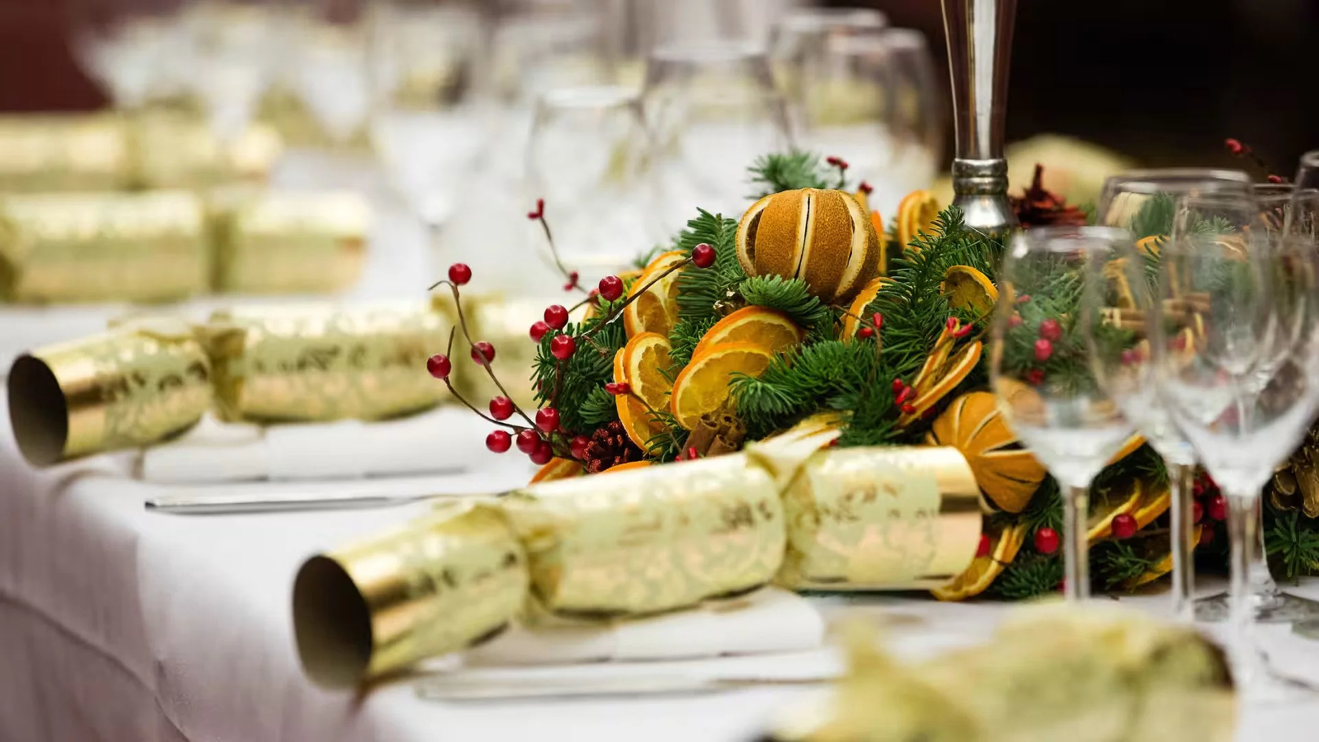 A table decorated for a celebration with a festive centerpiece of citrus slices, greenery, and red berries, surrounded by glasses and gold and white rolled napkins.