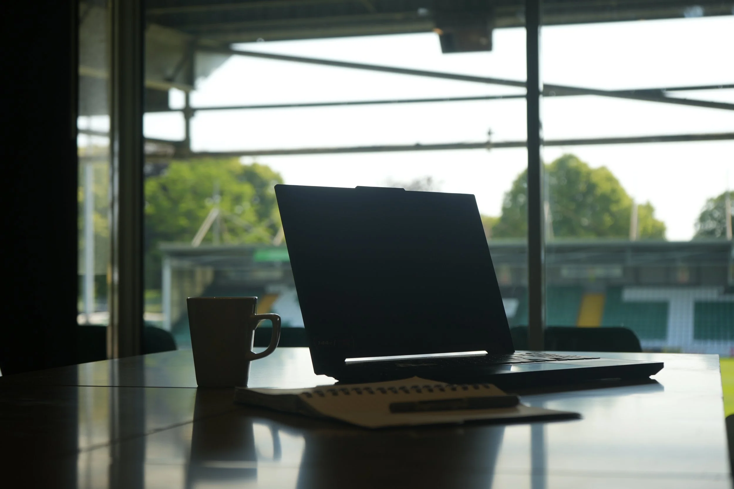 Laptop, mug, notebook, and pen on a table inside a room with large windows, showing an outdoor scene with trees and a sports field.