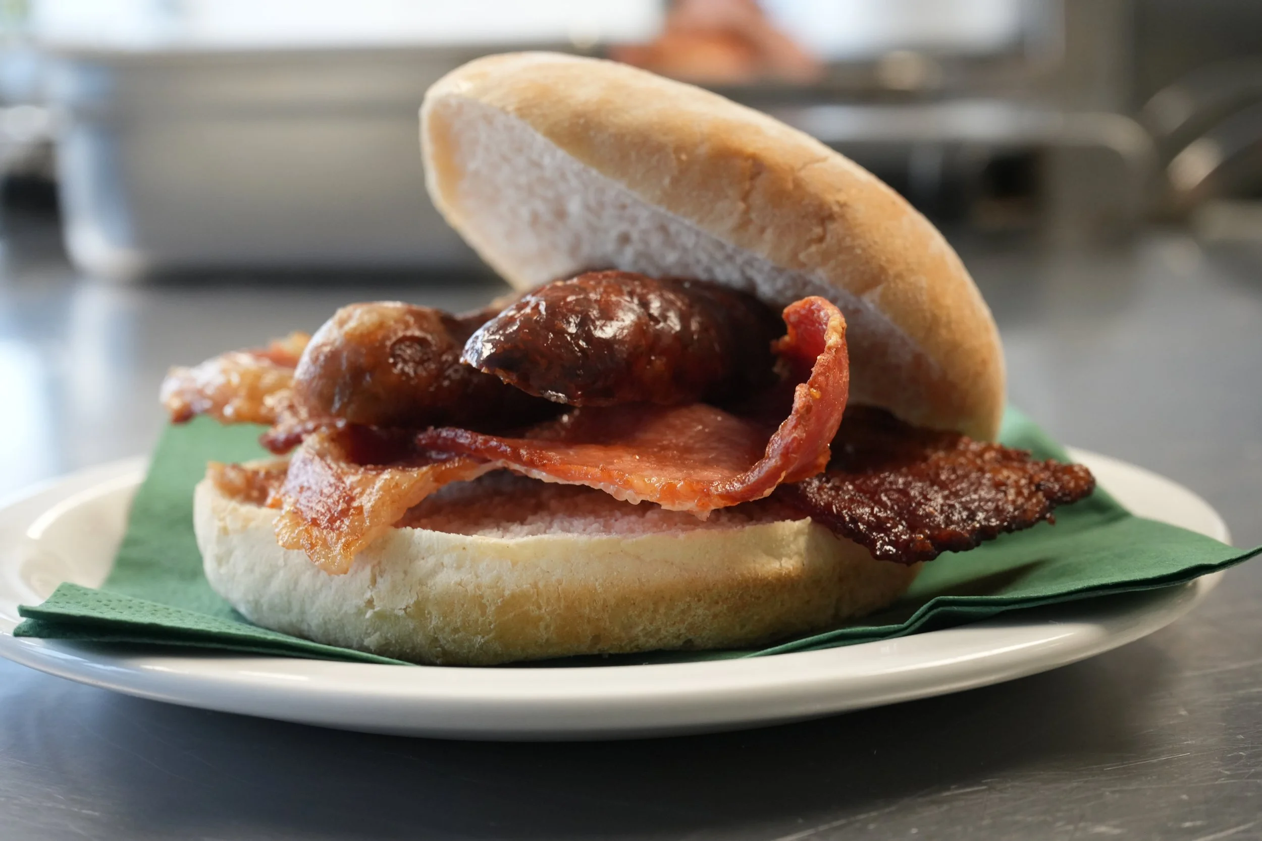 Close-up of a sandwich with bacon, sausage, and a bun on a plate with a green napkin.