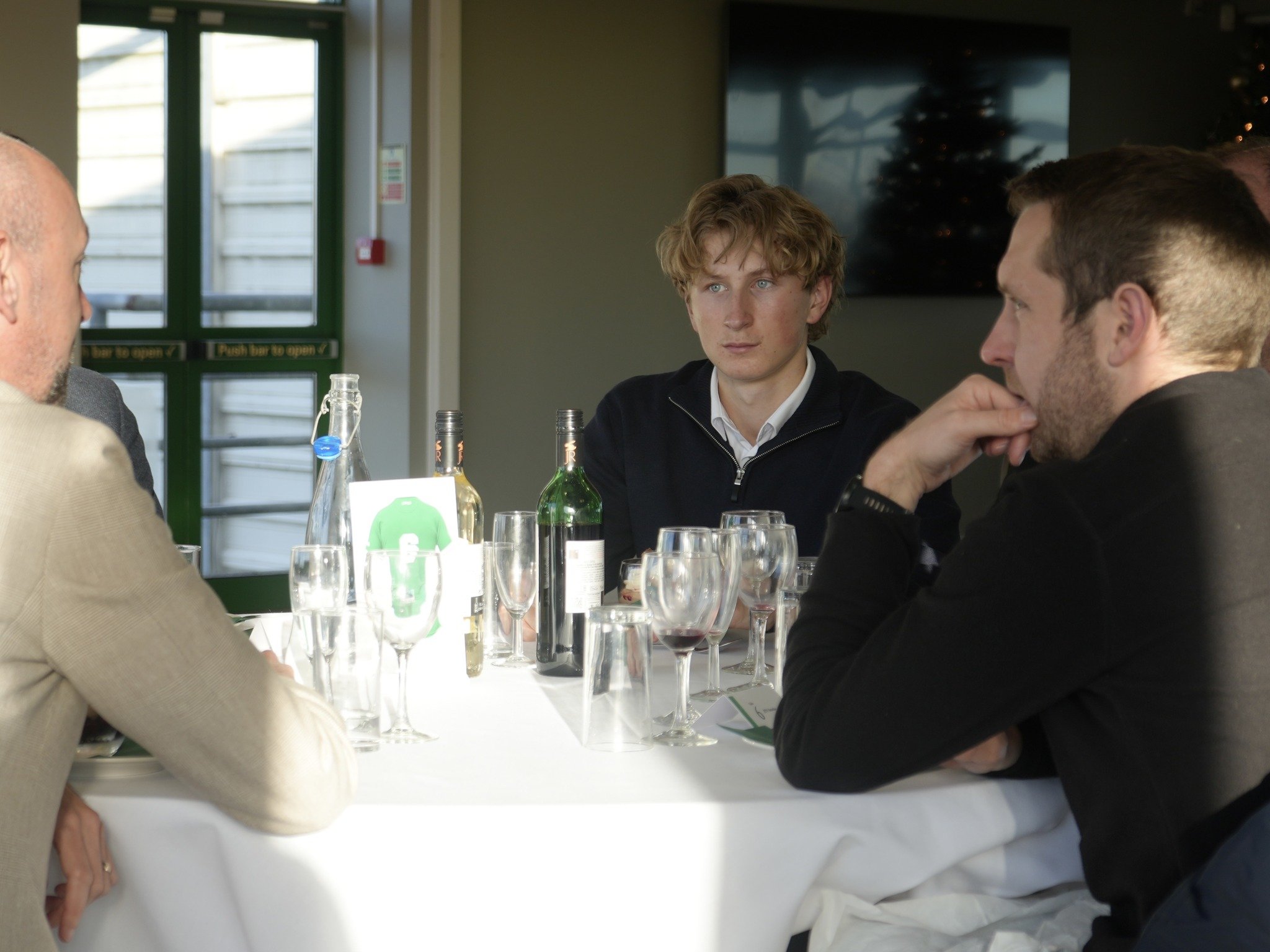 A group of men sitting at a table with empty glasses and bottles, engaged in a conversation, in a well-lit indoor setting with a television and Christmas tree in the background.