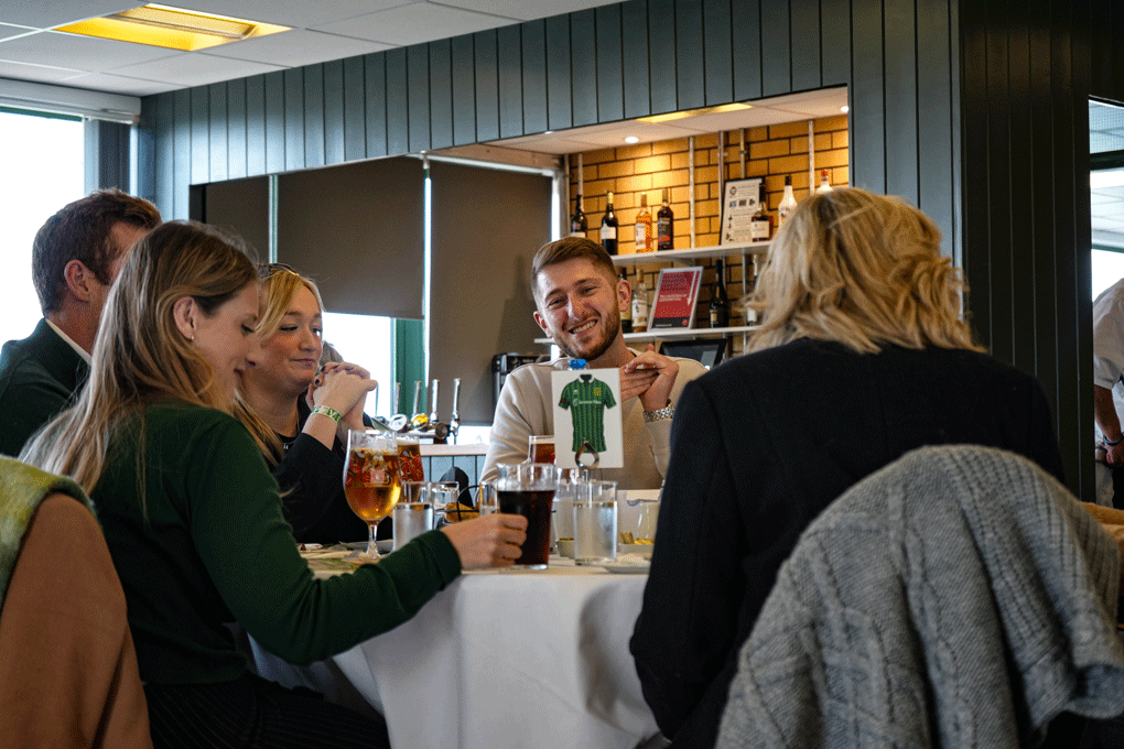 Group of six people sitting around a dining table in a restaurant, with a man holding up a green shirt-shaped object and smiling.