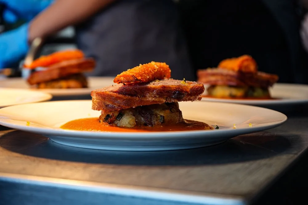 Close-up of a plated meal featuring a piece of fried pork belly on top of roasted vegetables with sauce on a white plate.