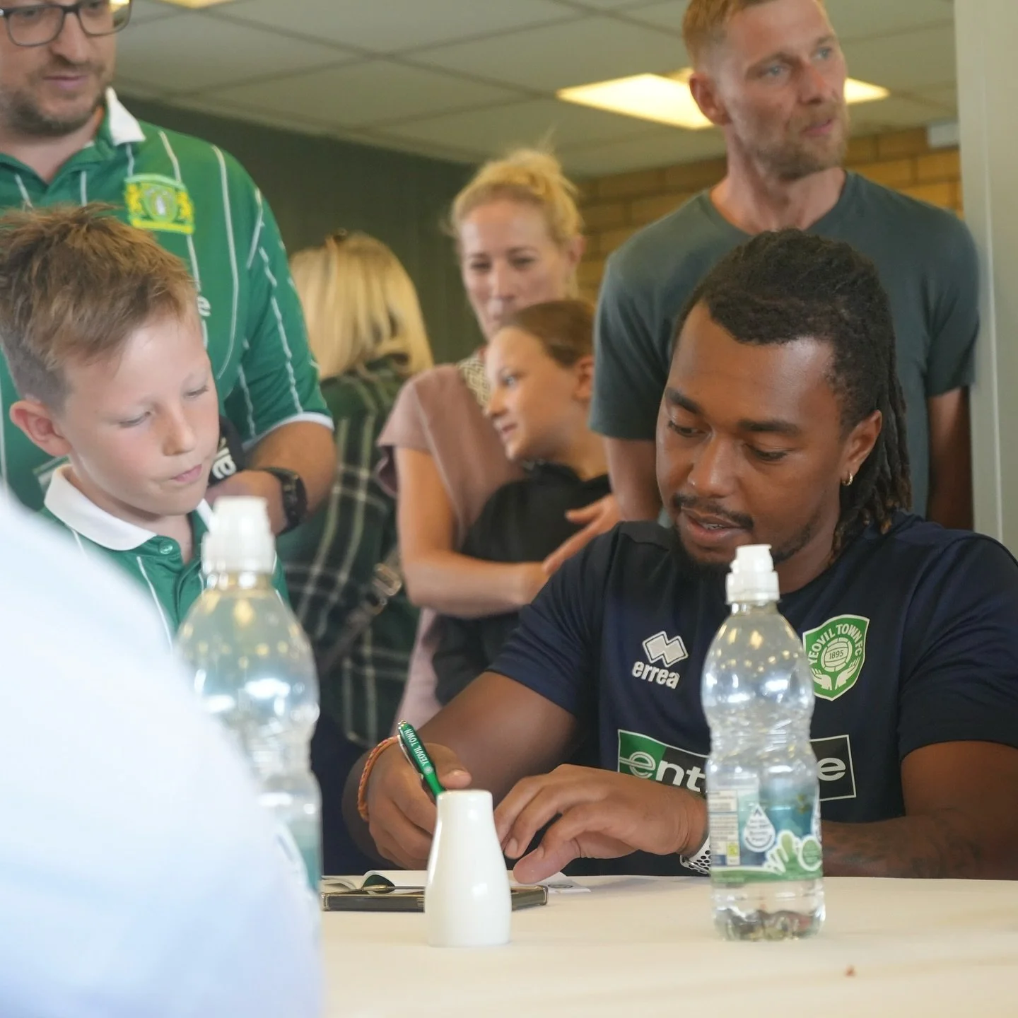 A man with dreadlocks, wearing a dark blue sports jersey with a green emblem, is signing an autograph at a table with water bottles and a small white object, while surrounded by young fans and other people in an indoor setting.