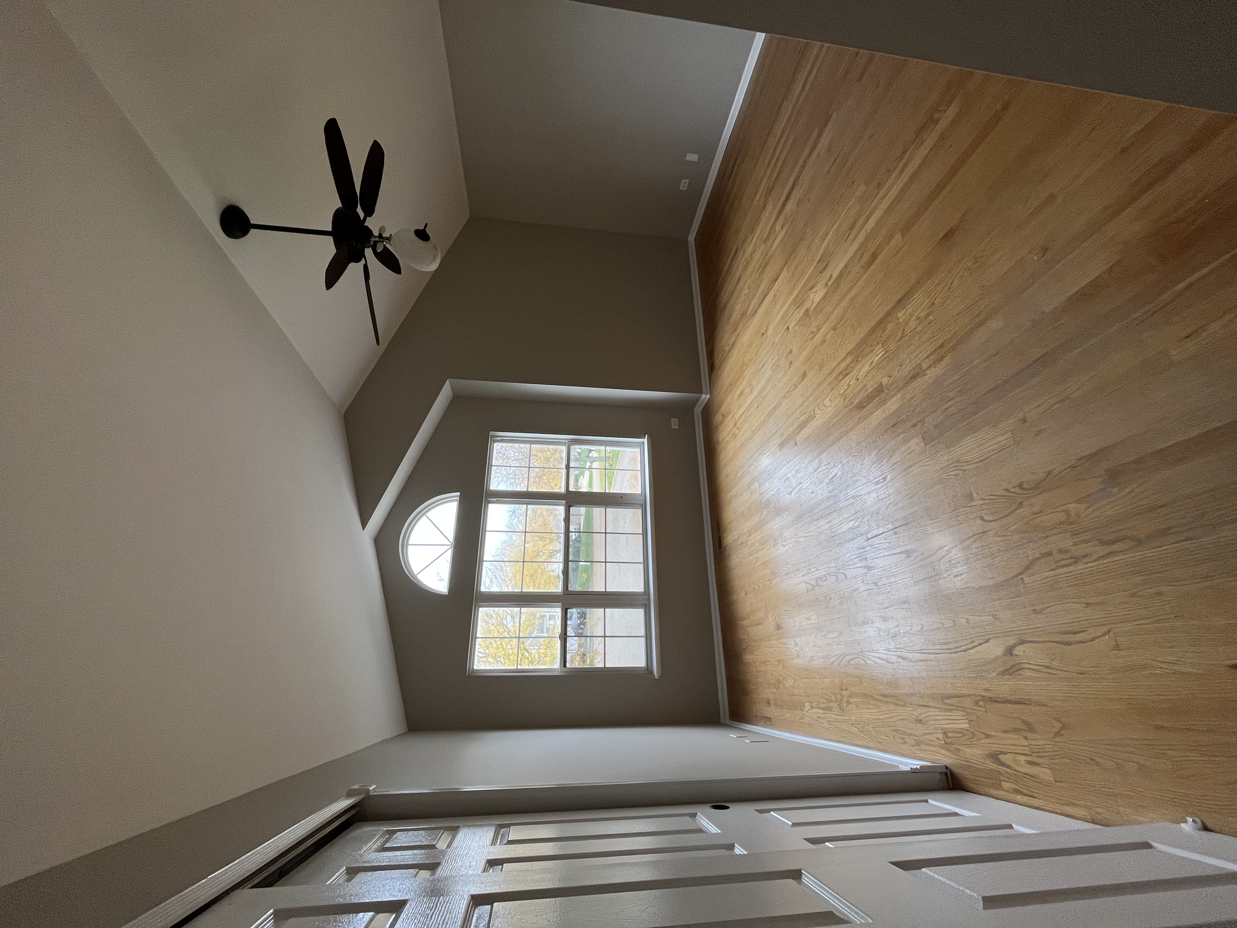 Interior view of an unfinished room with hardwood flooring, large windows, and a ceiling fan. The room is empty, with neutral wall colors and natural light coming through the windows.