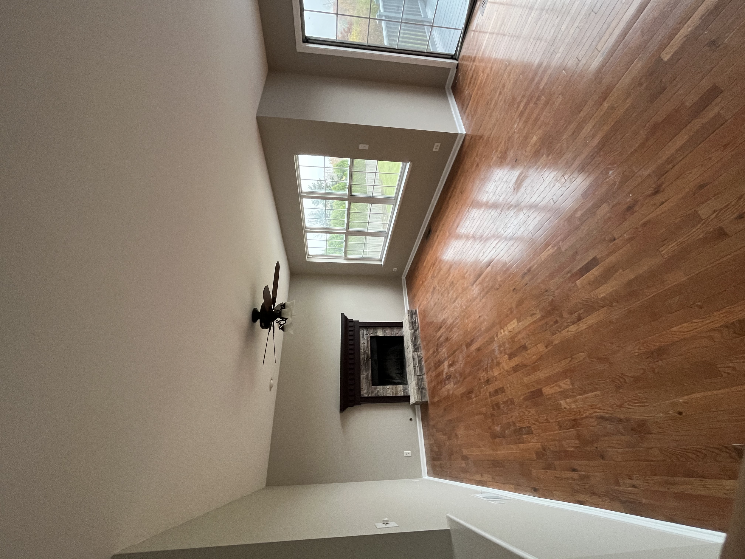 An empty living room with hardwood floors, a stone fireplace, large windows showing a green outdoor area, and beige walls.