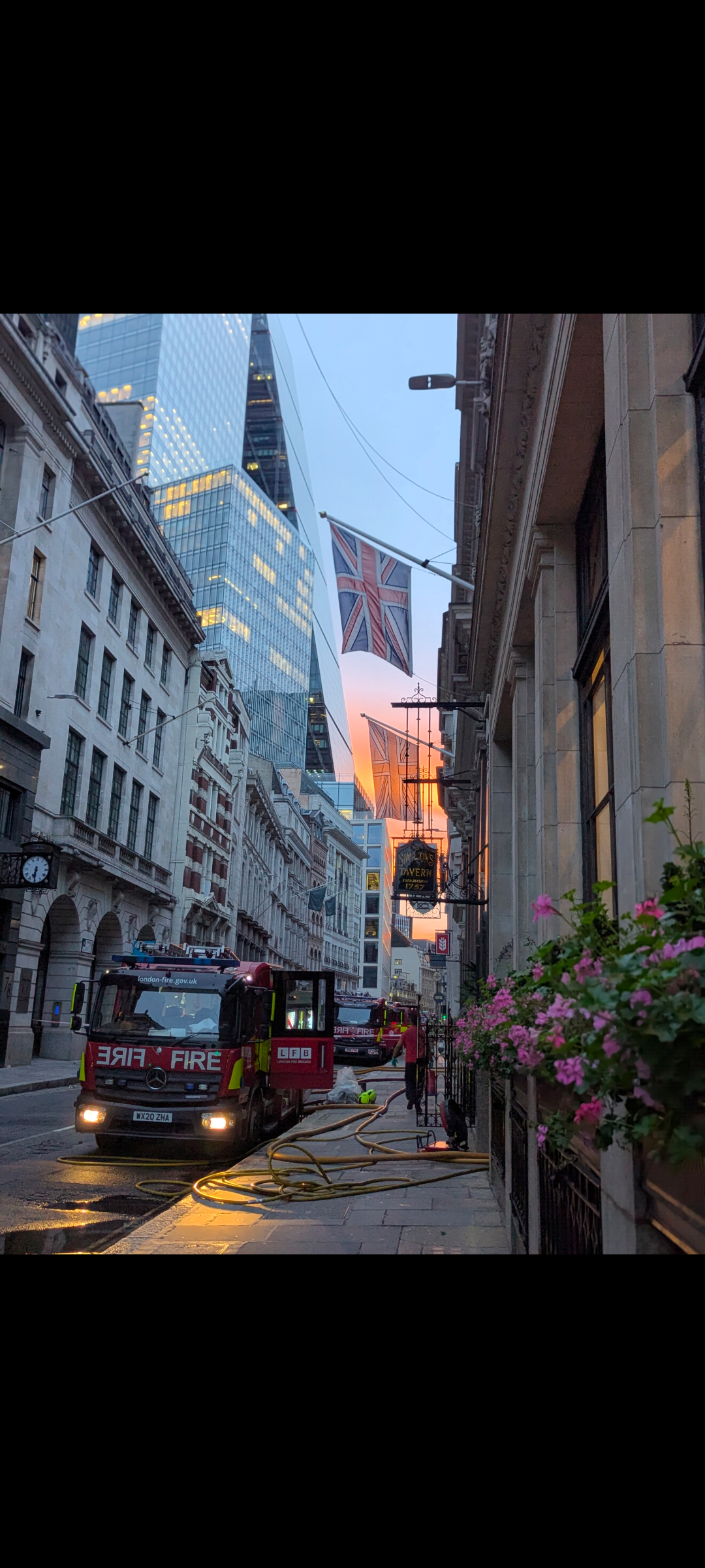 Fire trucks parked on a city street at sunset, with hoses on the ground and union Jack flags hanging between buildings.