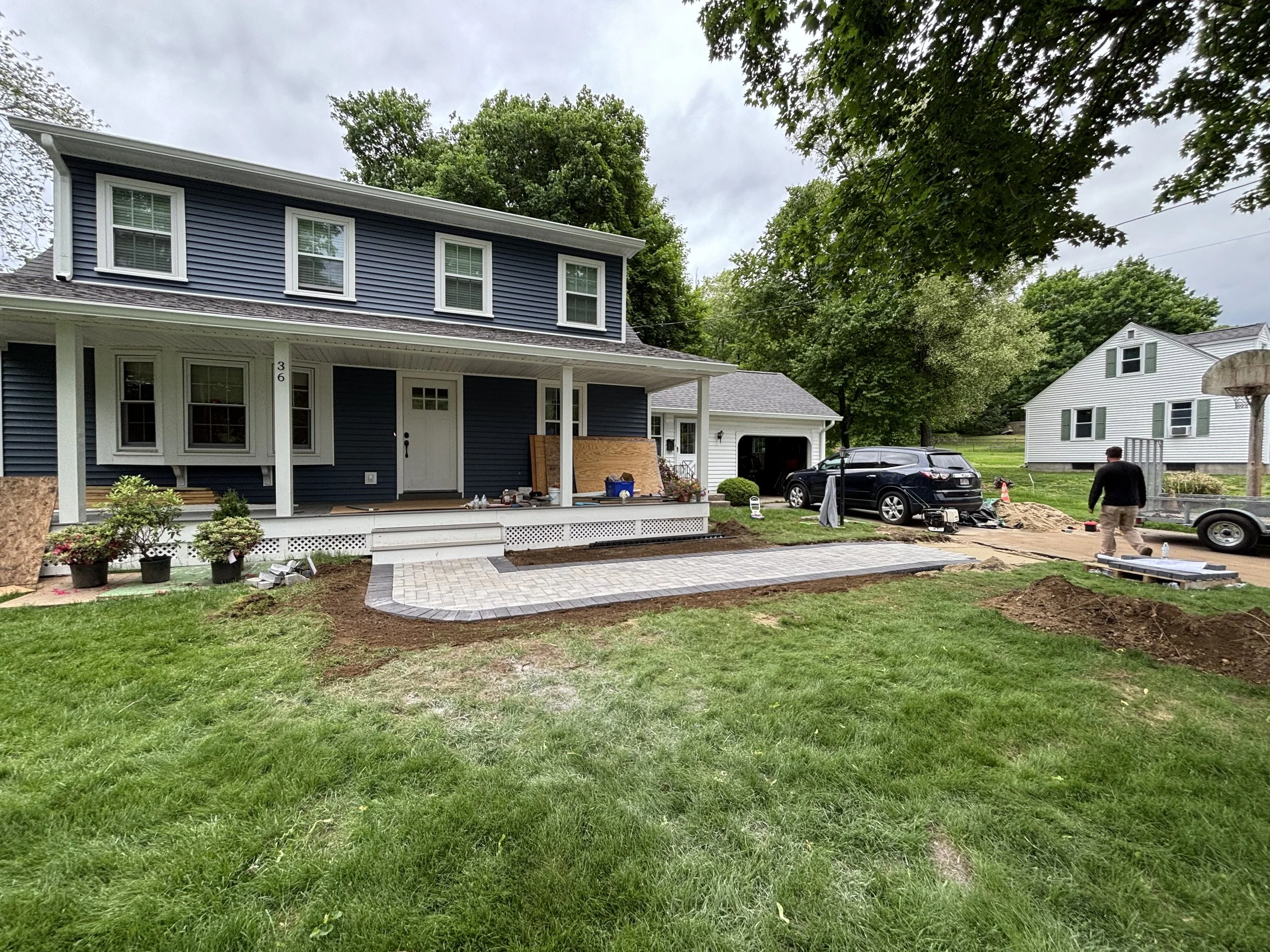 A house with a porch and a tilled yard under renovation. There are two men working, a black SUV, potted plants, and construction materials. The yard has a partial newly paved pathway.