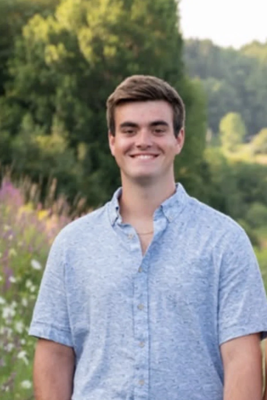 A smiling young man with short brown hair, wearing a light blue button-up shirt, standing outdoors in a lush green park with trees and flowers in the background during daylight.