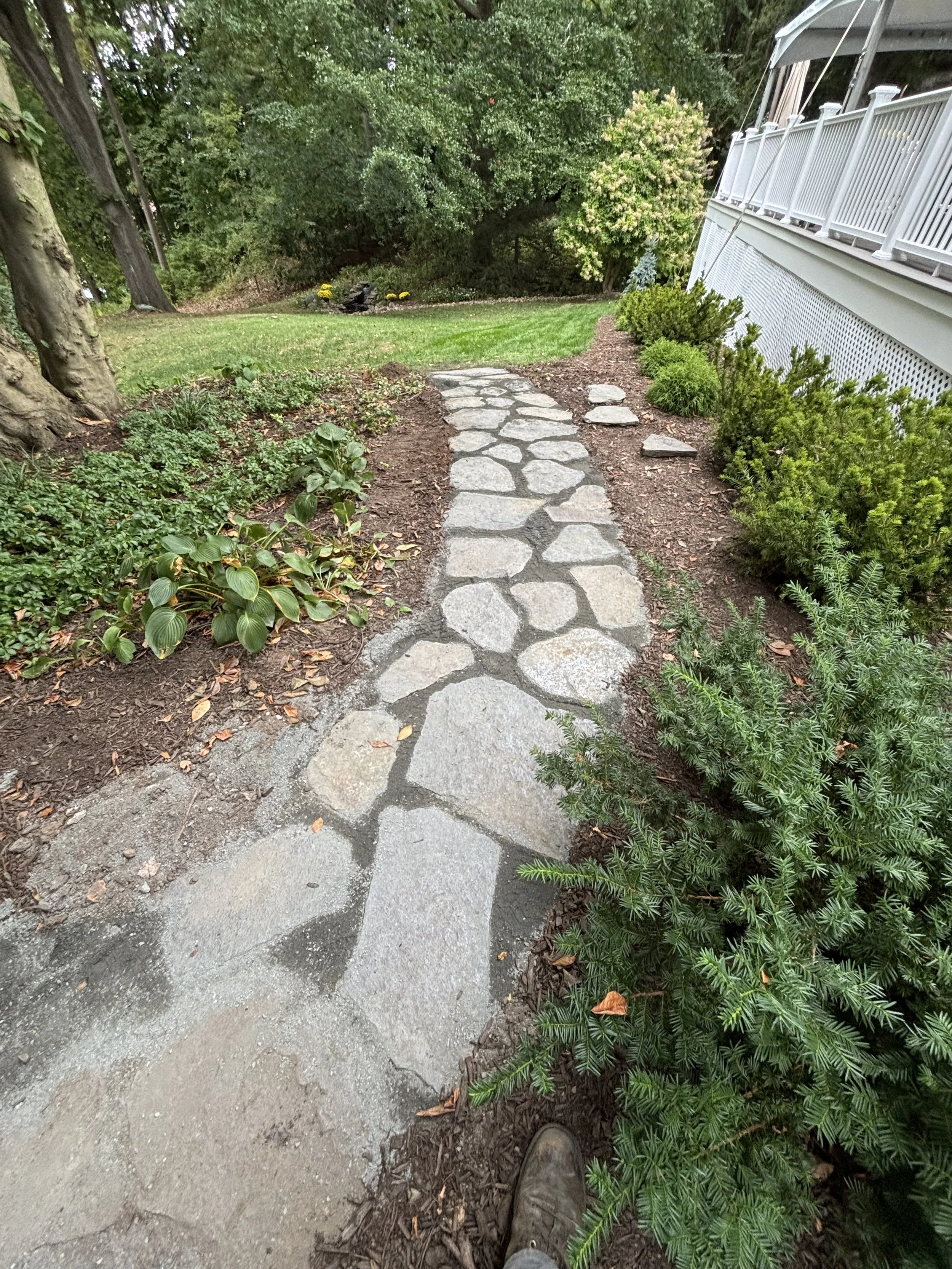 Stone pathway bordered by green bushes and plants, leading through a garden with trees and grass, with a white porch railing on the right side.