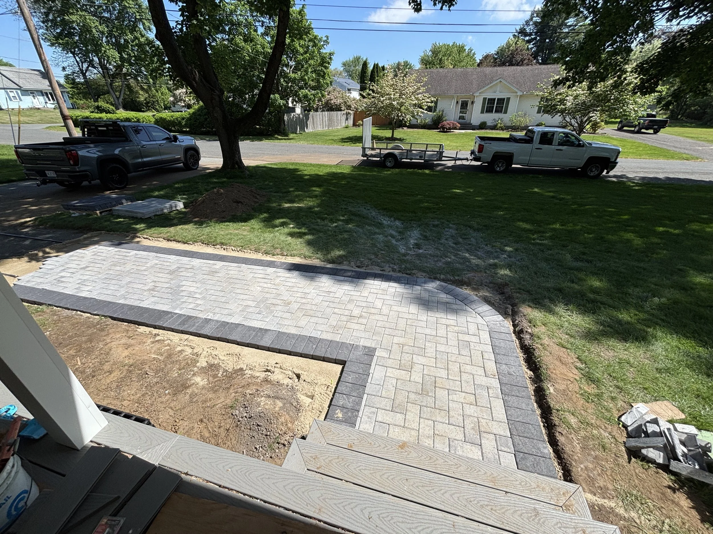 View from a porch showing a newly paved brick walkway leading to a grassy yard, with trucks parked on the street and trees in a suburban neighborhood.