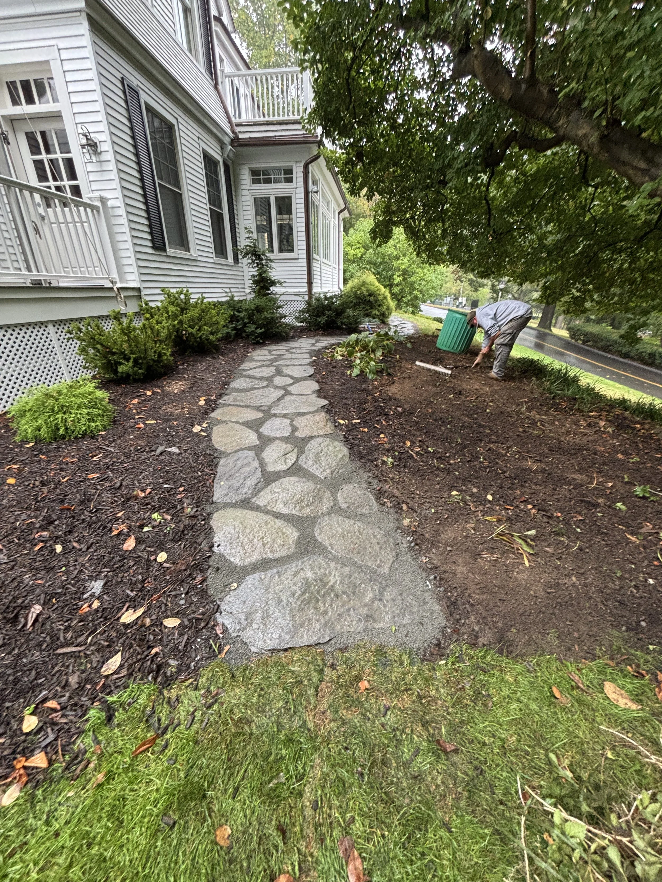 A man working on planting or gardening beside a house with a stone pathway, surrounded by green bushes, trees, and recently tilled soil.