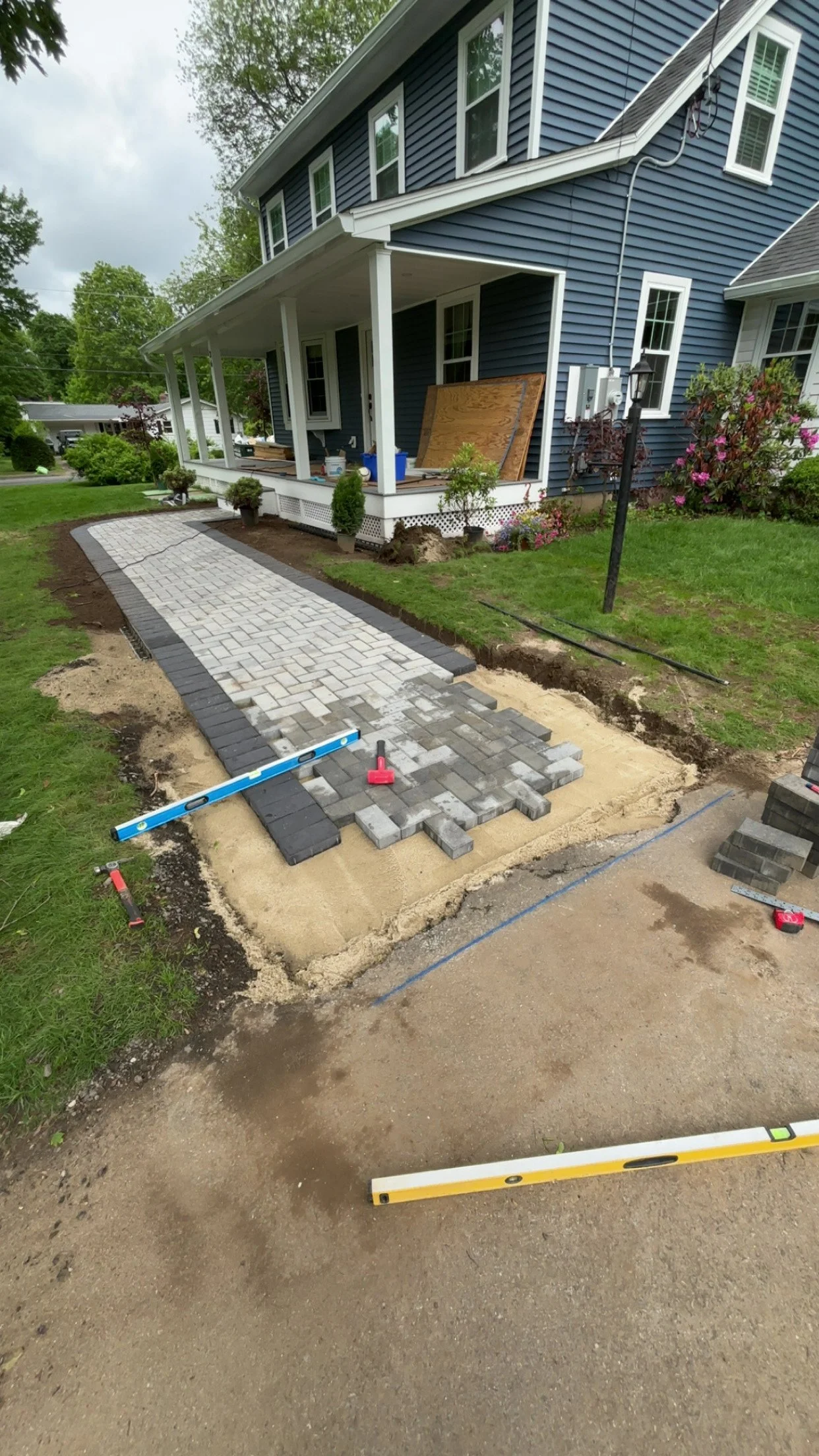 Installing a brick walkway in front of a blue house with a porch. Some bricks are already laid, and tools are visible near the pathway.